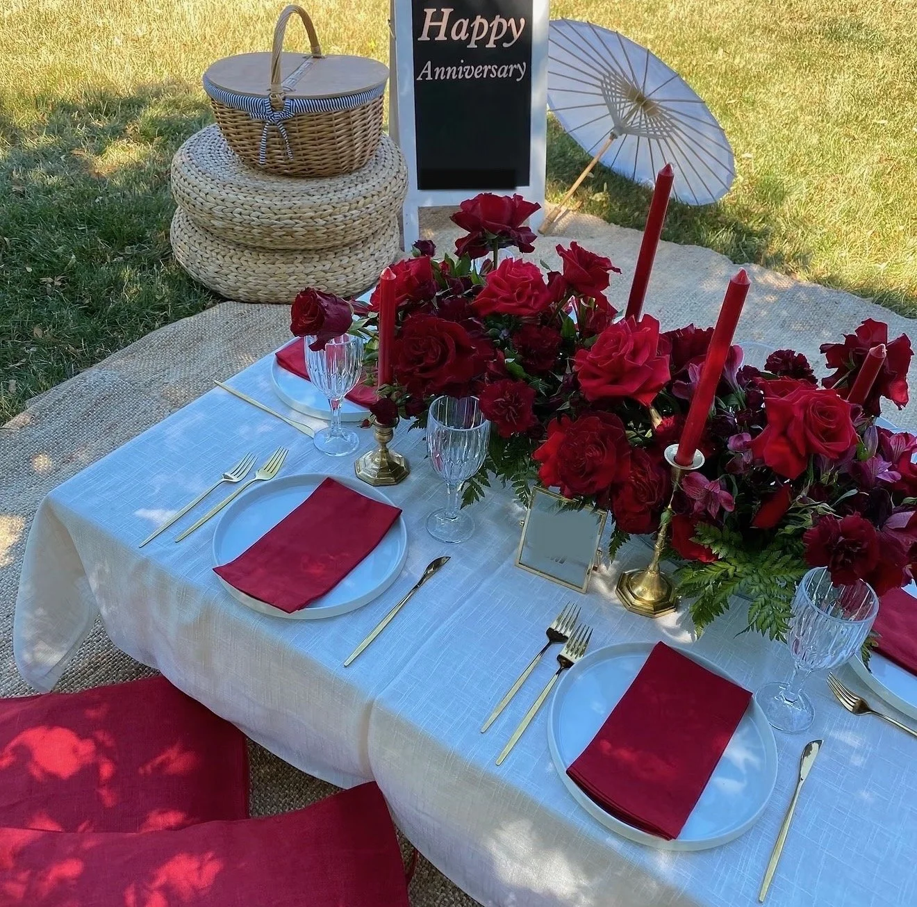 Romantic outdoor anniversary picnic featuring red roses, red taper candles, white plates, gold cutlery, crystal glassware, and a Happy Anniversary sign on a grassy lawn.