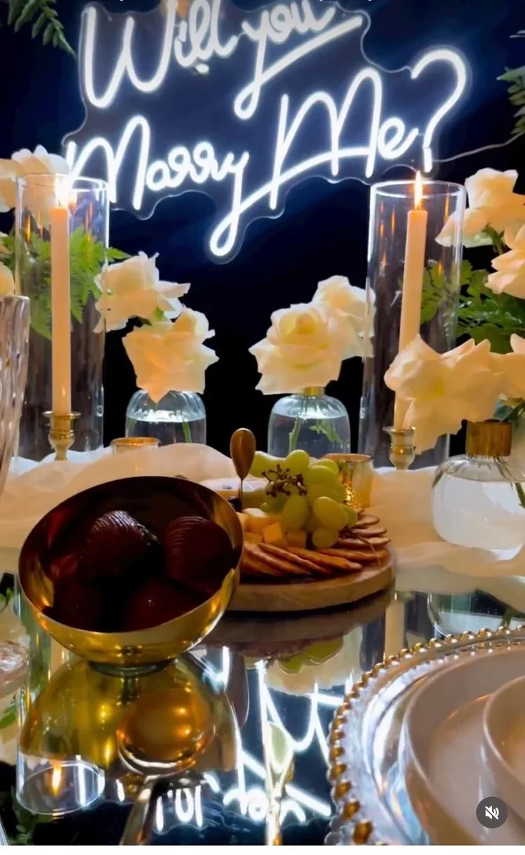 A festive table setting featuring a glow-in-the-dark neon sign that says "Will you marry me?" surrounded by white flowers, glass candle holders with lit candles, a cheese platter, grapes, and various tablescape decorations.