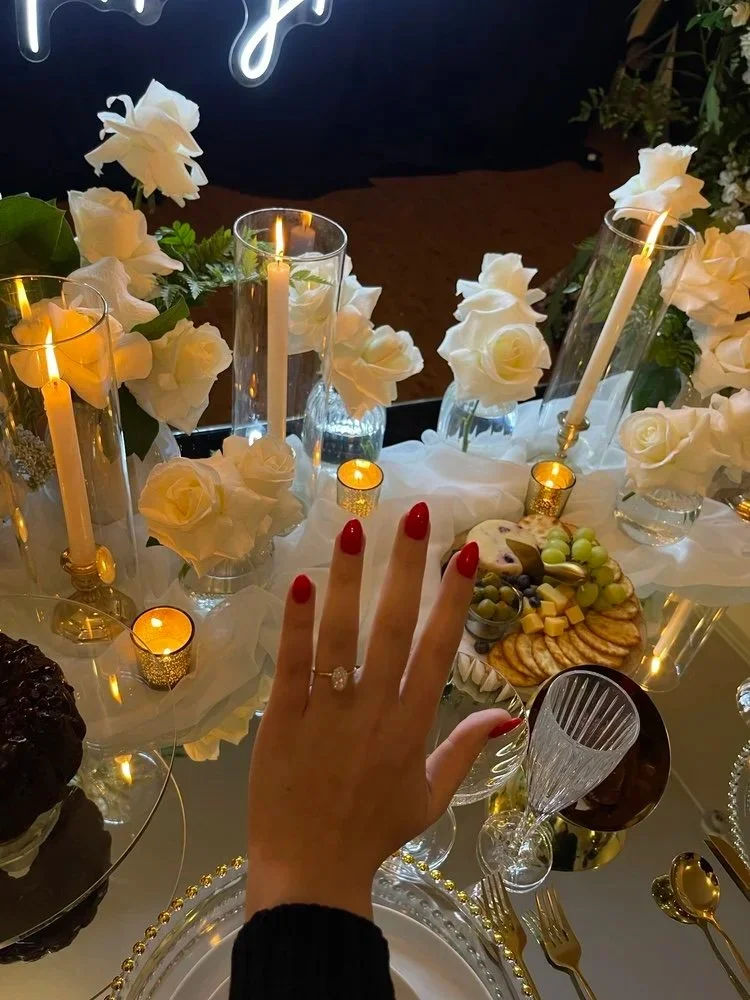 A person's hand with a ring and red nail polish is displayed over a decorated dining table with white roses, tall candles in glass holders, small candles, and a variety of snacks and desserts.