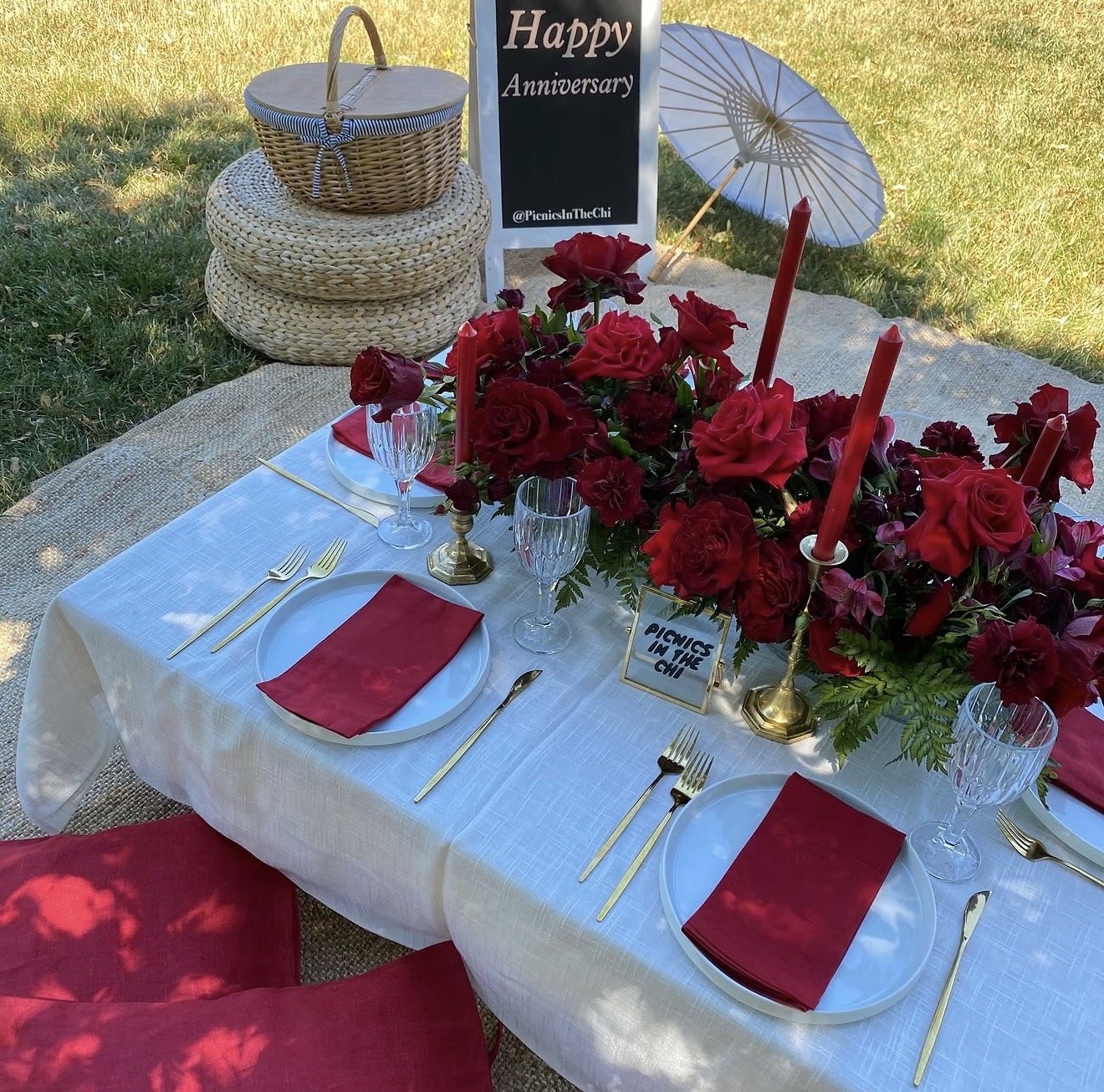 Romantic outdoor anniversary picnic featuring red roses, red taper candles, white plates, gold cutlery, crystal glassware, a wicker basket, and a Happy Anniversary sign on a grassy lawn