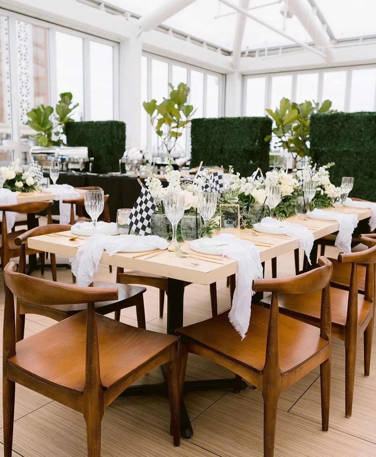  Elegant celebration dinner setup with long wooden tables, white floral arrangements, checkered flags, crystal glassware, and wooden chairs in a bright indoor venue.