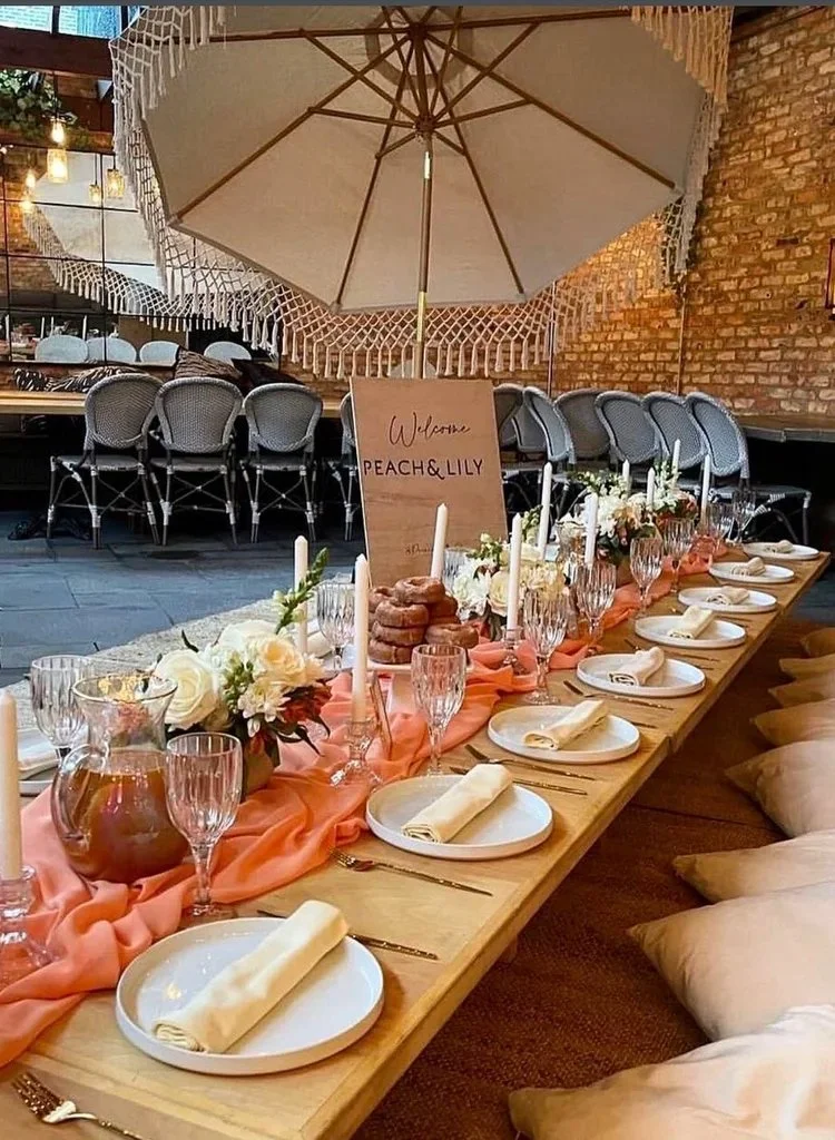 Indoor dining setup with a long wooden table decorated with peach-colored fabric, white flowers, candles, and tableware including plates, napkins, and glasses. In the background, a brick wall and a sign that reads 'Welcome Peach & Lily' are visible, 