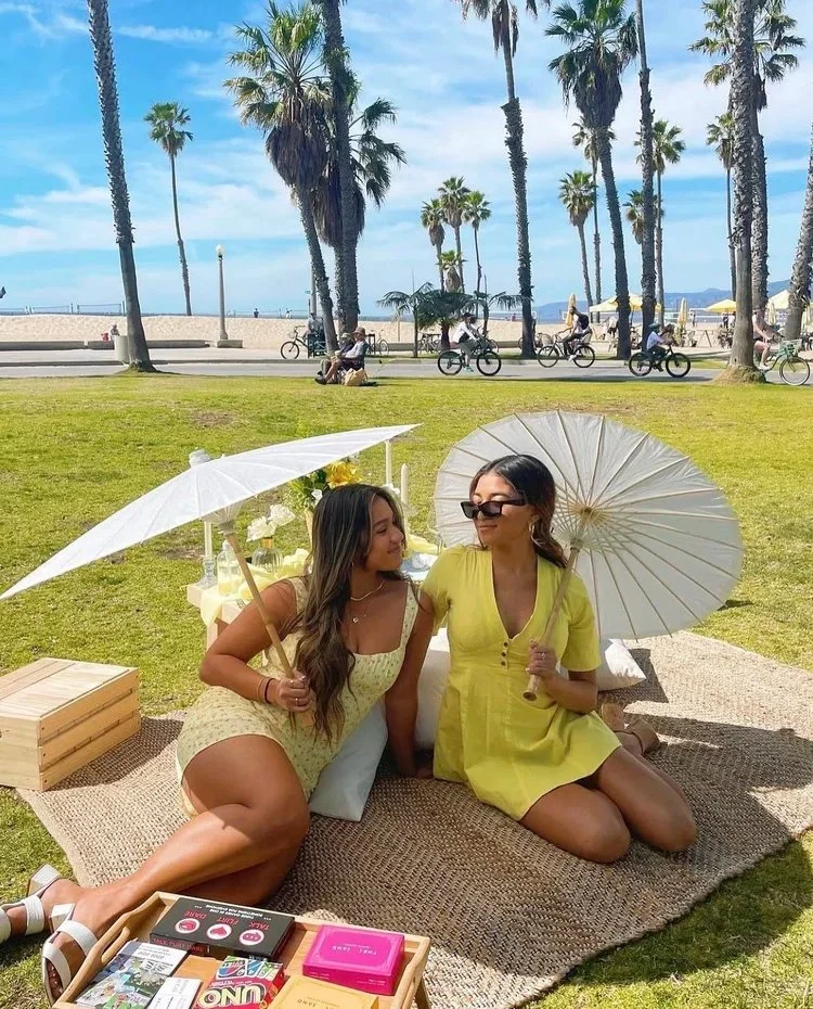 Two women sitting on a woven blanket at a park, holding white paper parasols, with a table of colorful books in front of them, and palm trees in the background on a sunny day.