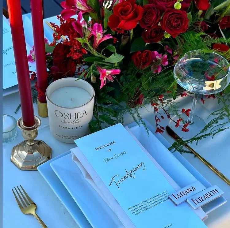 A table decorated with red and pink flowers, a white candle labeled Oshea Candles with fresh linen scent, a gold candlestick with two red candles, a clear glass bowl with water and flowers, a white plate with a blue napkin, a gold fork, a gold knife,