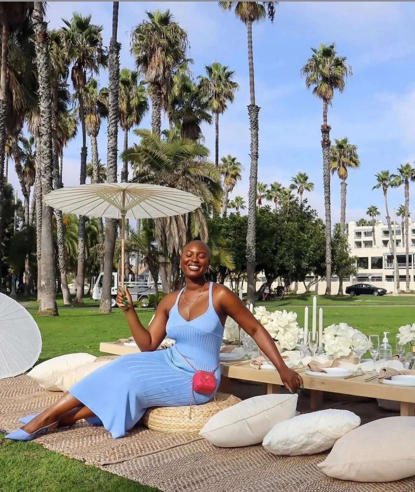 Woman in a blue dress enjoying a luxury outdoor picnic with elegant tableware, white floral arrangements, and plush cushions under a sun umbrella.
