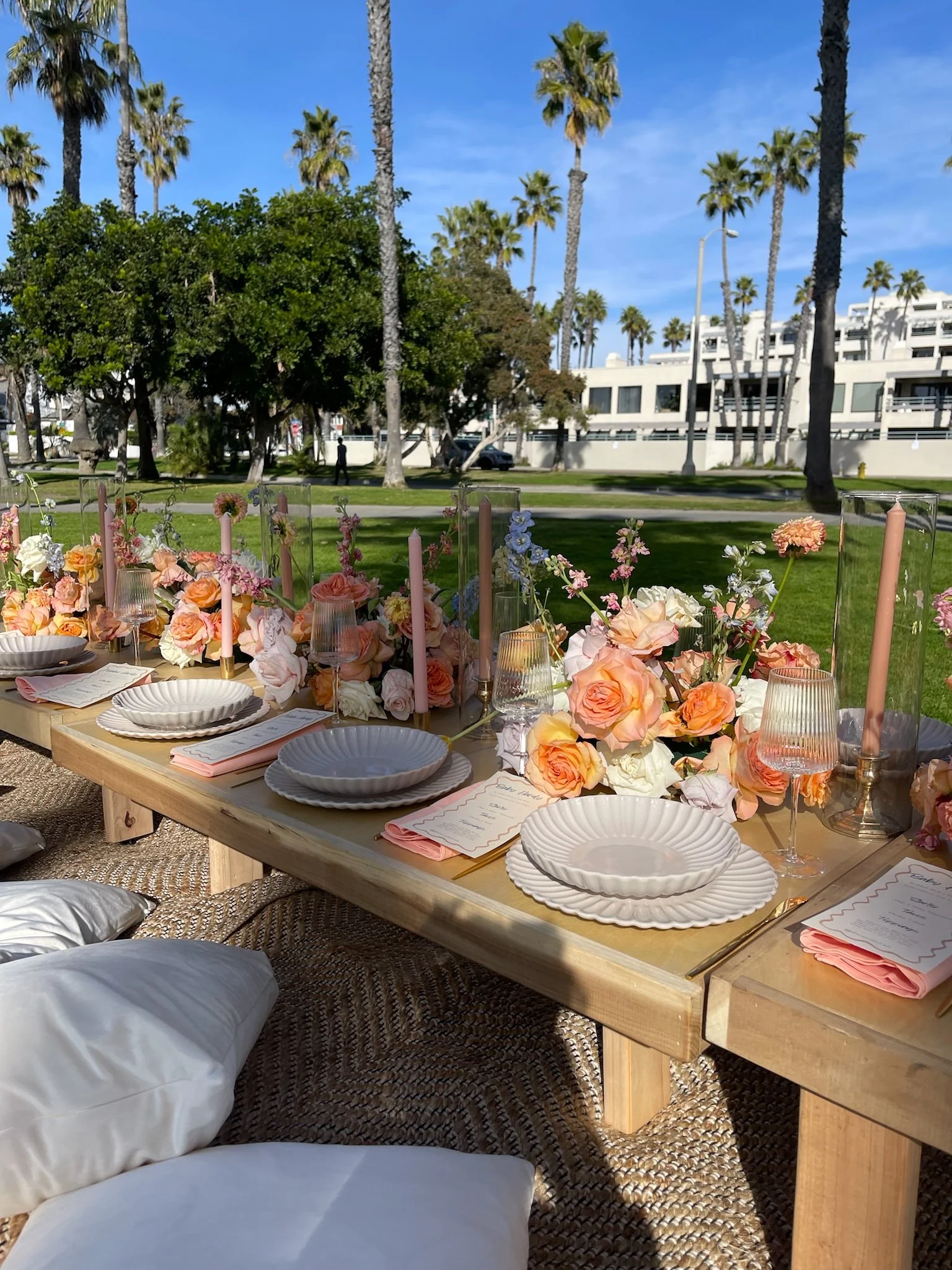 Luxury outdoor picnic tablescape with peach roses, elegant white plates, glass vases, decorative menus, and woven rugs beneath palm trees.