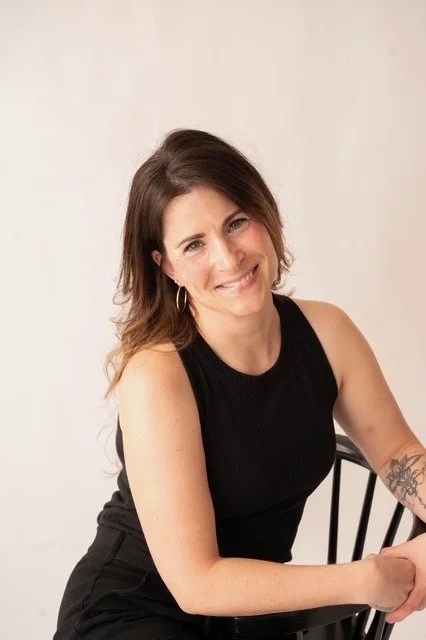 A woman with brown wavy hair, wearing black glasses and a black shirt, smiling at the camera with a floral tattoo on her left forearm, standing against a white background with geometric patterns.