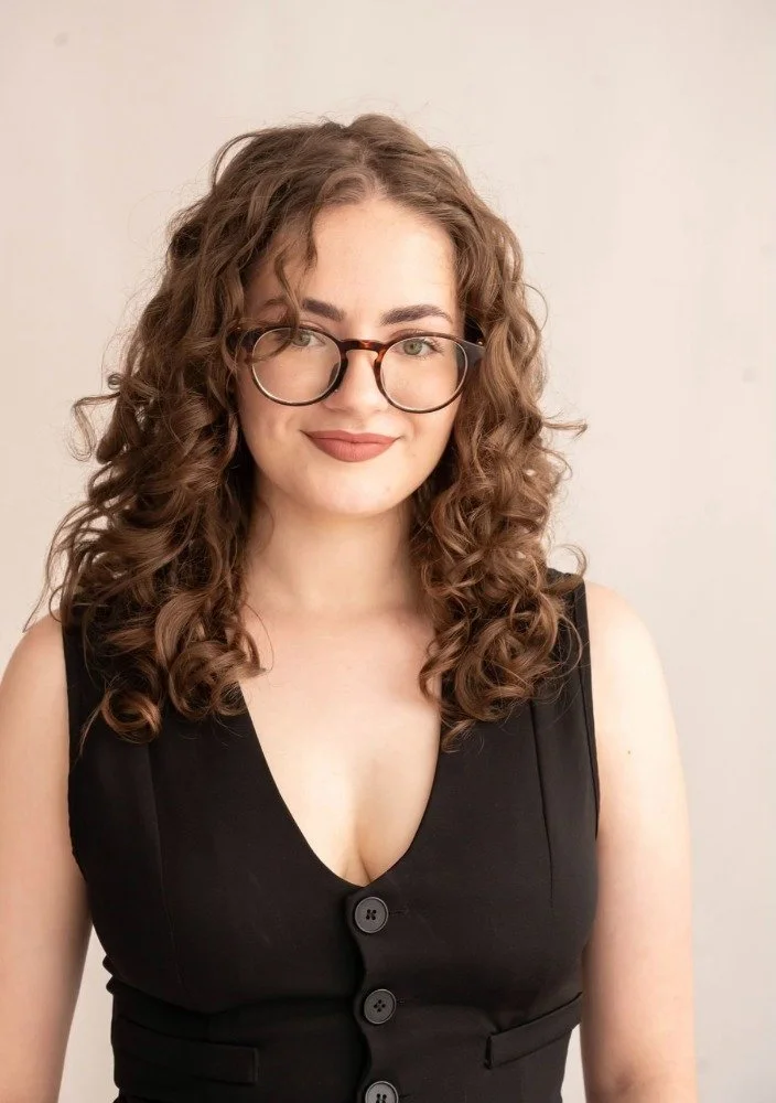 Woman with curly brown hair wearing a beige sleeveless top and white pants, standing crossed arms, smiling, against a white geometric wall background.