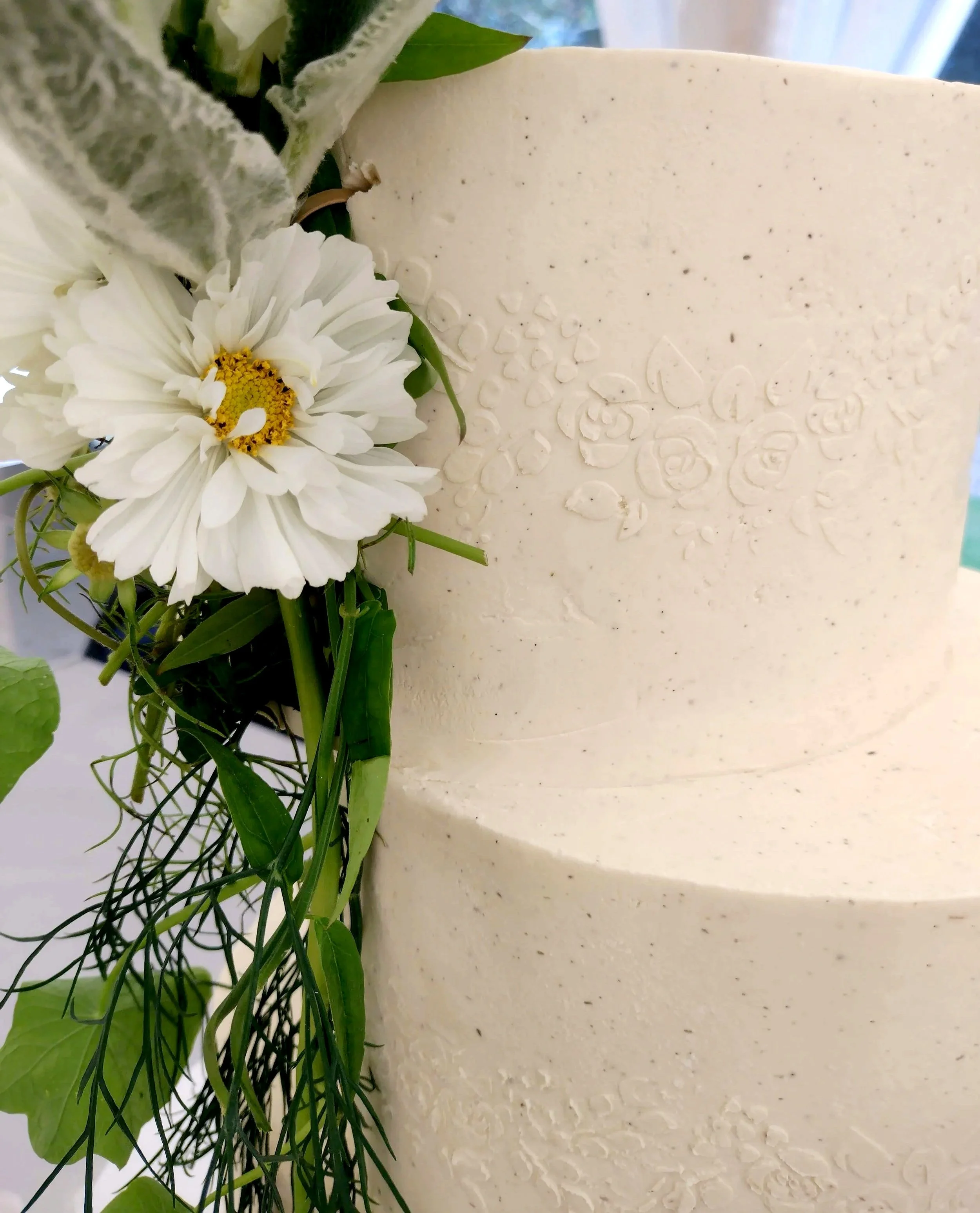 Relief stencil work of a floral rose wreath in white chocolate ganache lead your eye to the posy of seasonal, organic white and green flowers and foliage which adorned this bespoke wedding cake. *Due to the heat in the marquee the wedding co-ordinato