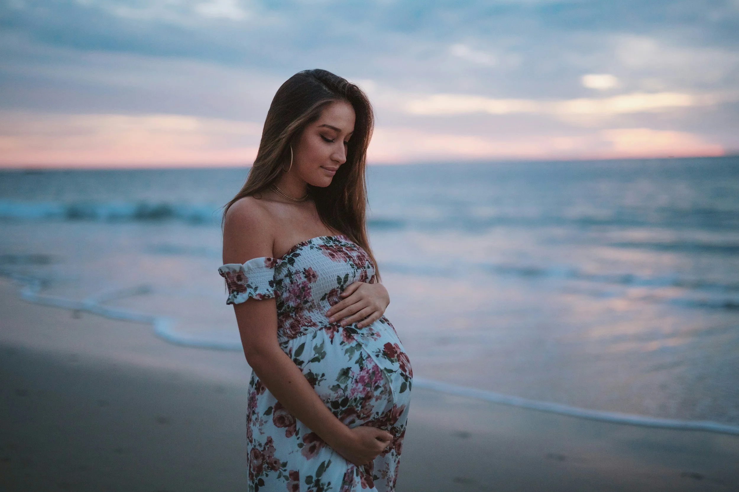 Pregnant woman standing on the beach at sunset with hands on belly, preparing for postpartum support.