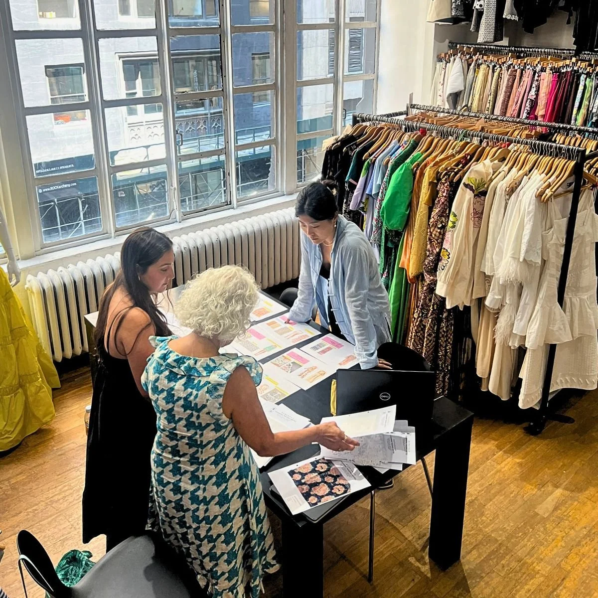 People examining documents and fabric swatches in a brightly lit fashion studio, surrounded by racks of clothing.