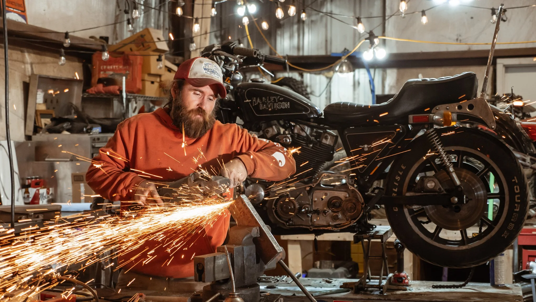 Bearded man grinding metal with sparks flying, standing in front of a Harley-Davidson motorcycle – rugged industrial photography with dramatic lighting.