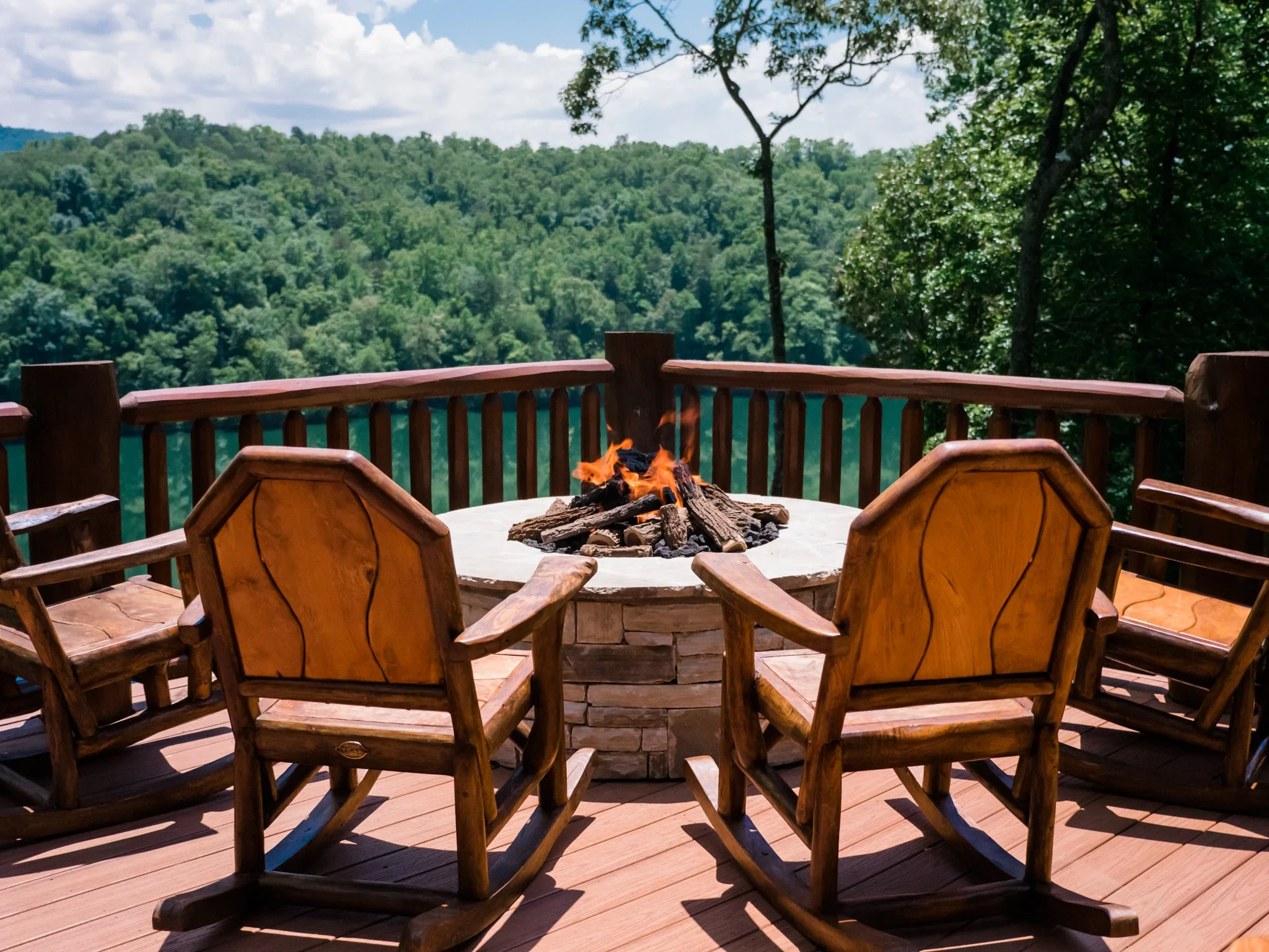 Wooden rocking chairs around an outdoor fireplace on a balcony, overlooking a serene lake in the mountains – cozy and scenic mountain retreat setting.