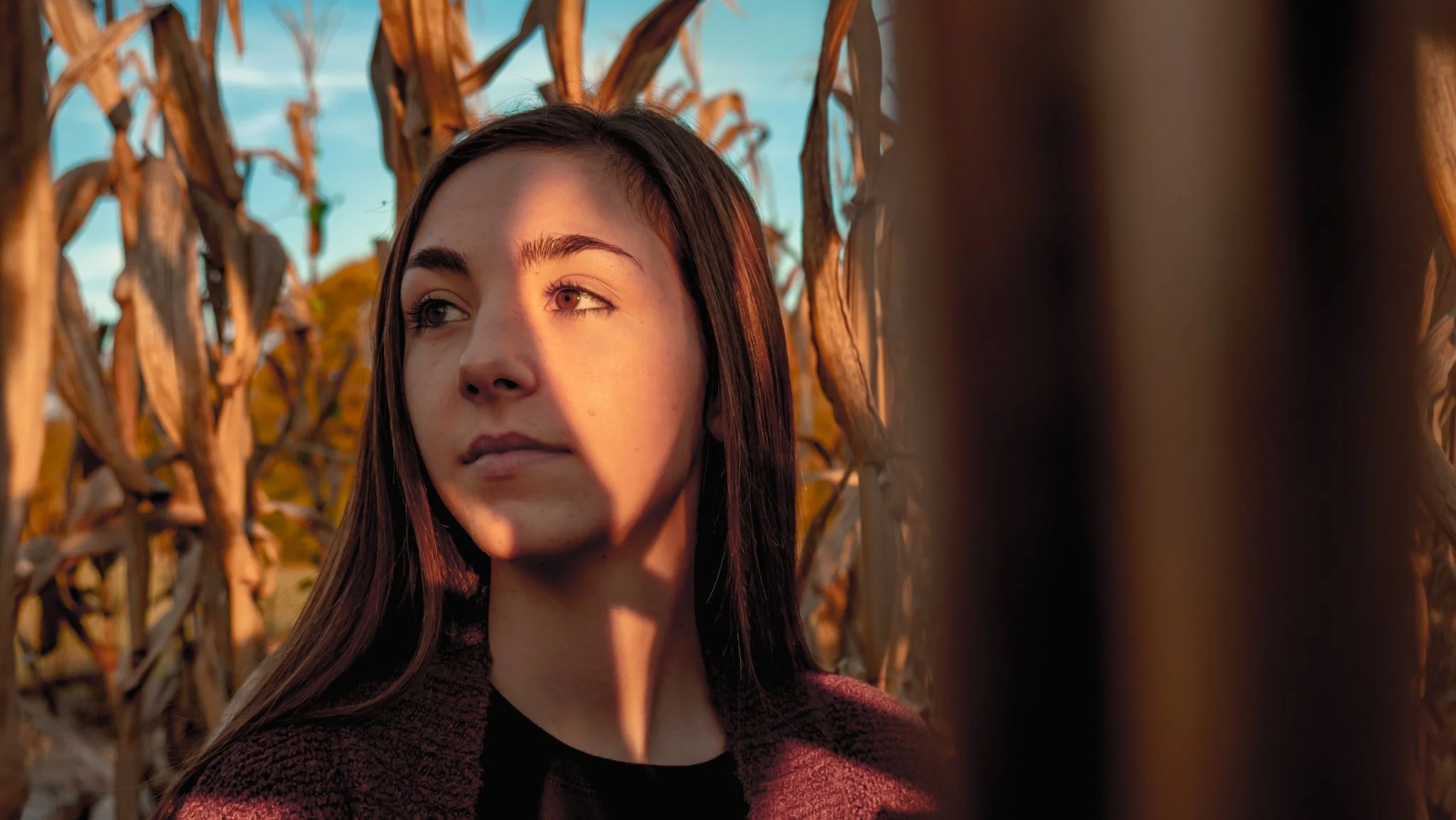 Young girl standing in a cornfield, smiling and looking away as the warm sunset light gently illuminates her face – serene and joyful outdoor portrait.