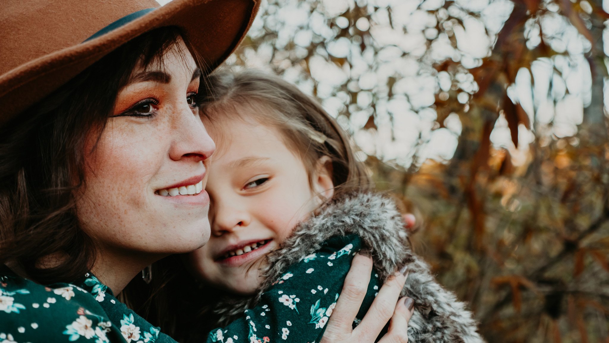 Mom and little girl wearing stylish clothes, holding each other while smiling and gazing off into the distance – heartwarming outdoor family portrait