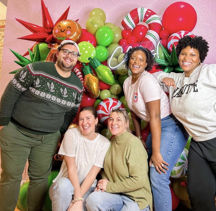 Five people posing in front of a red and green Christmas balloon wall with Christmas themed mylar balloons.
