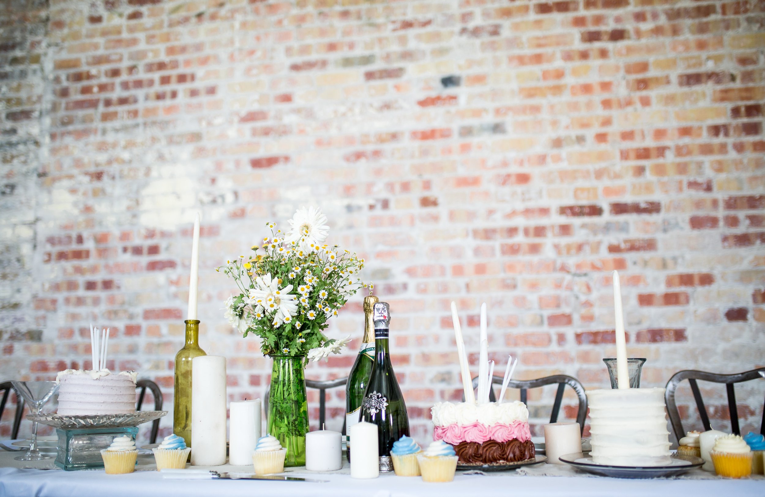 A spread of desserts, candles, and flowers on a table with metal chairs behind it as well as an exposed brick wall.