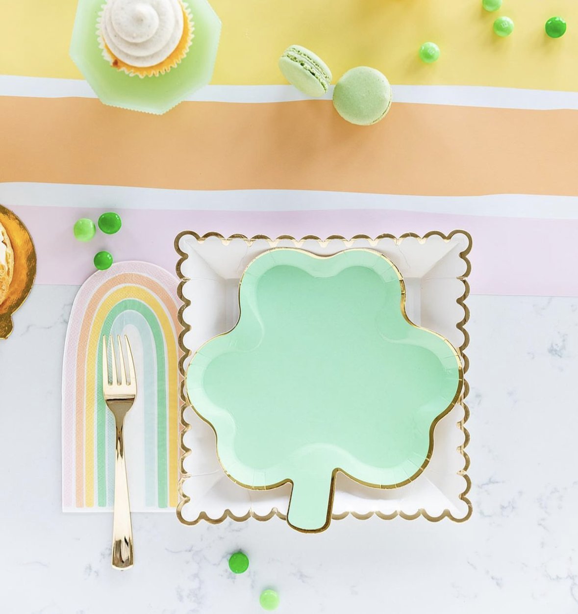 An overhead shot of a table with a green shamrock plate, a gold lined scalloped plate, a rainbow napkin, and gold fork.