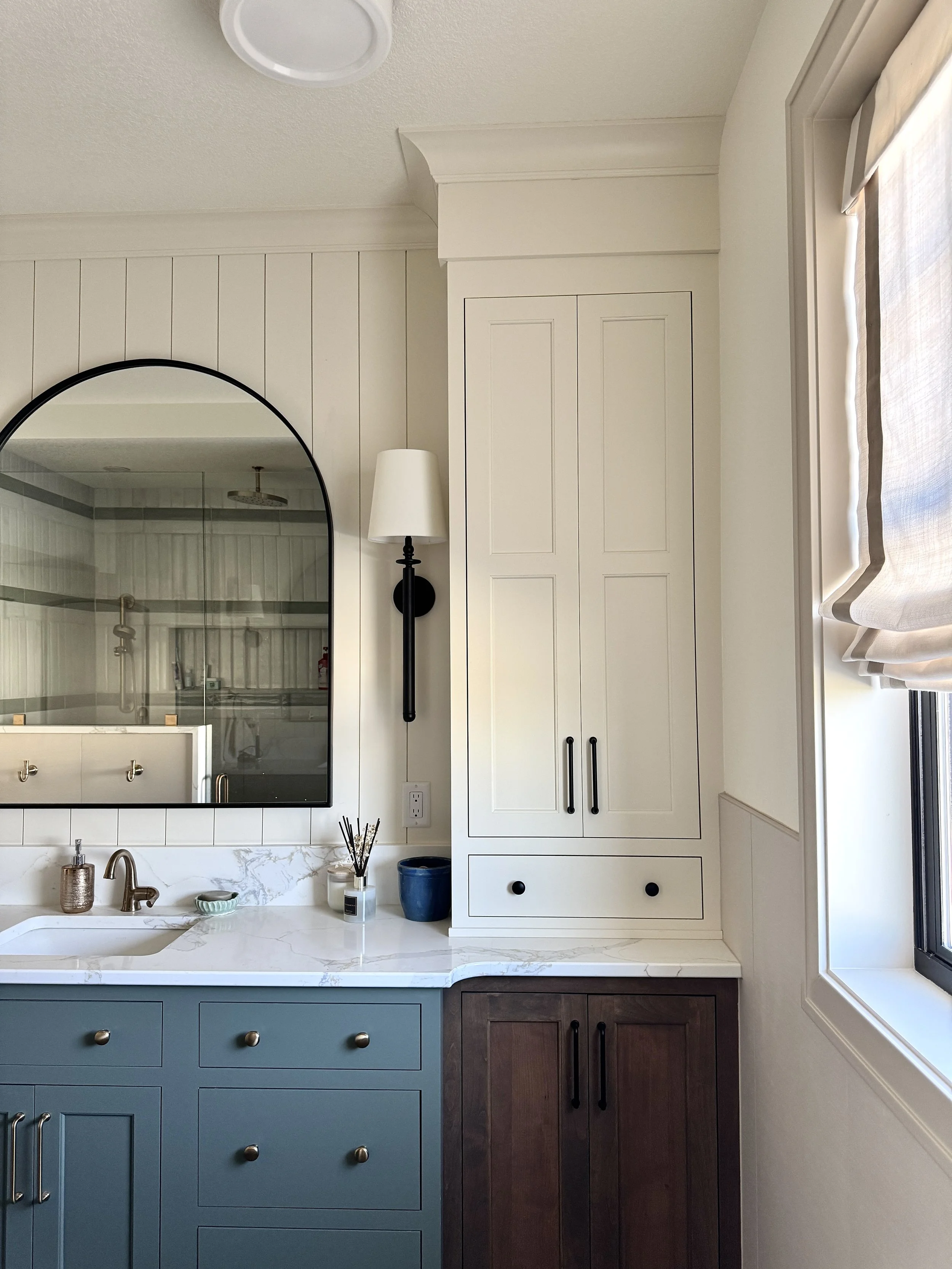 A bathroom vanity with a white marble countertop, a blue drawer cabinet, and a dark wooden cabinet, a large mirror, a wall-mounted light, and a window with blinds.