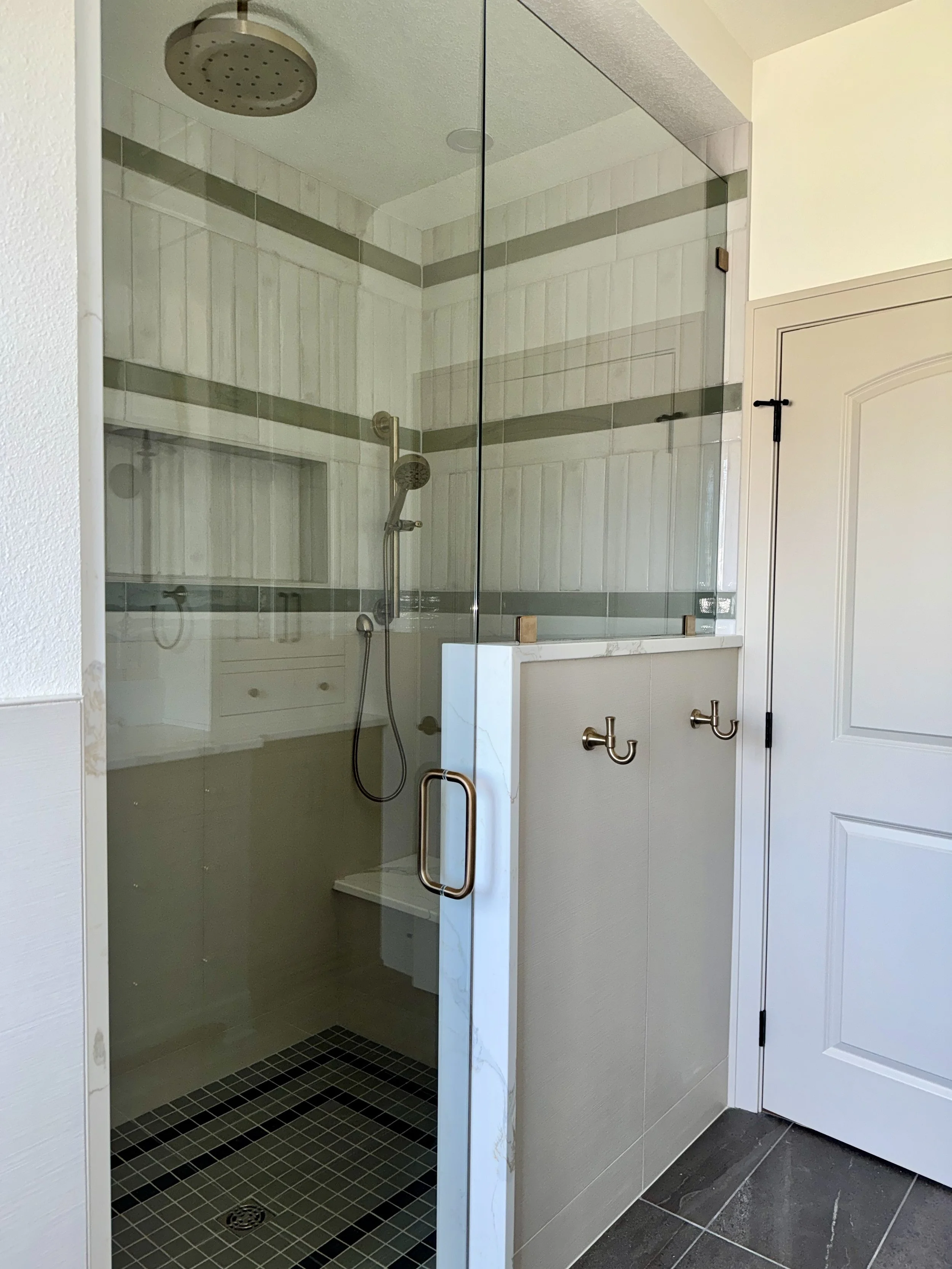A glass-enclosed shower with a rain shower head on the ceiling and a handheld shower head on the wall, featuring white wooden wall tiles with gray horizontal stripes, a marble-topped half-wall with hooks, and a door with black hinges and handle in a 