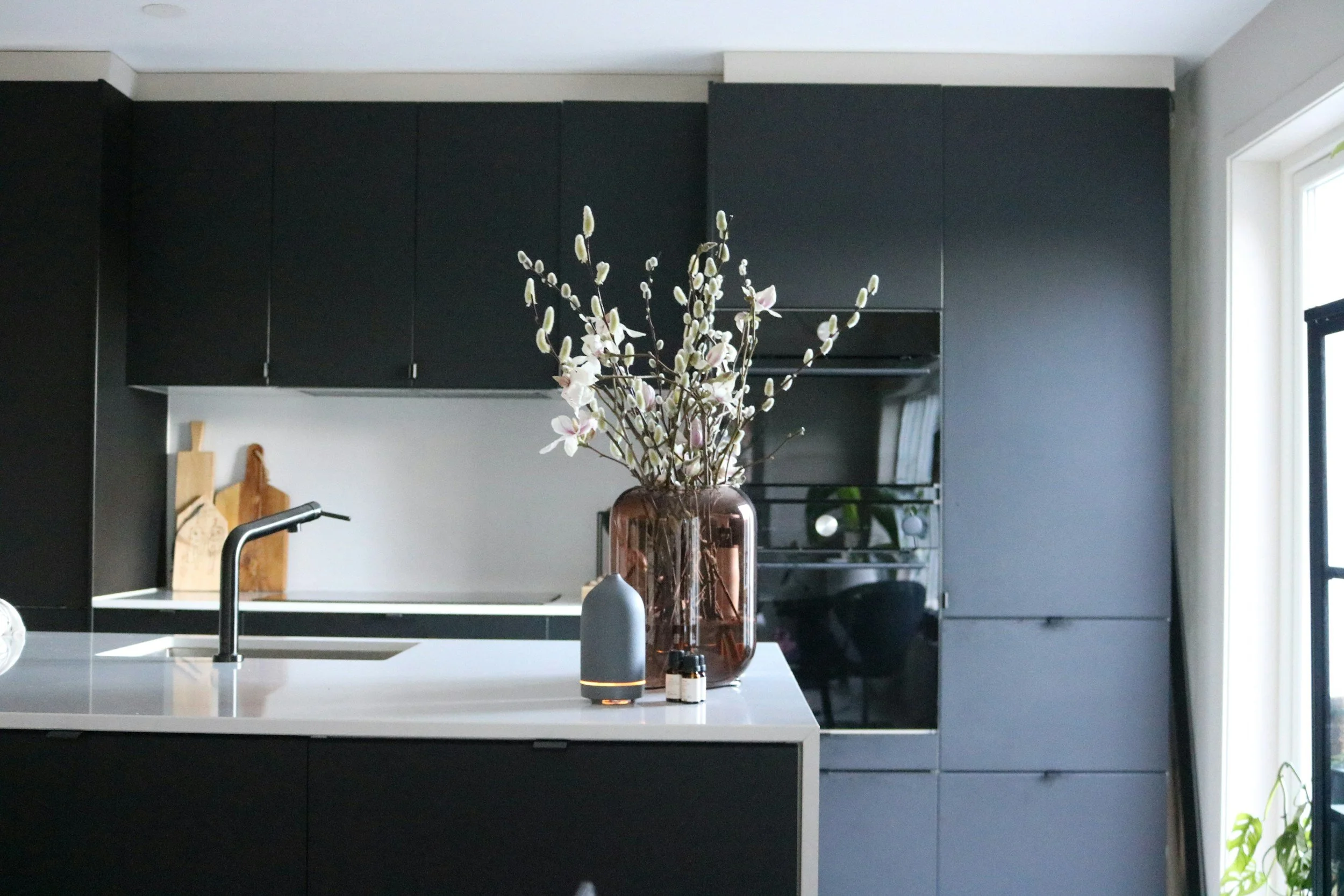 Modern kitchen with black cabinets, white countertop, central faucet, large vase with white flowers and pussy willow branches, and a window on the right side.