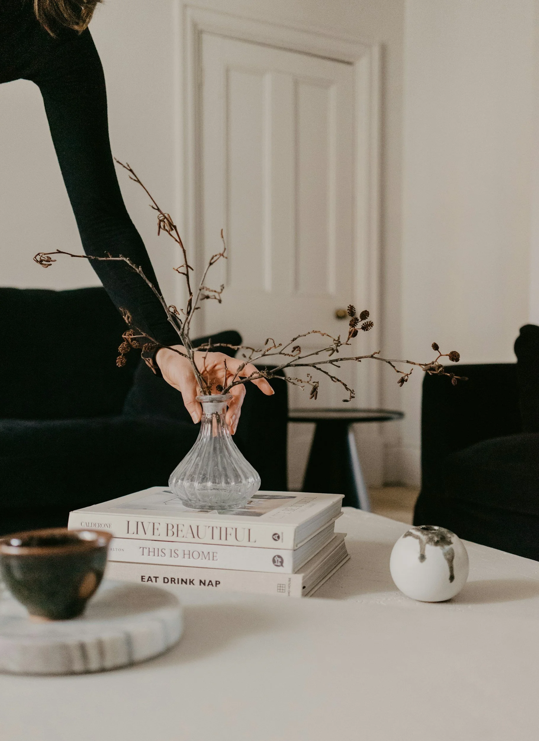 A person arranging dried branches in a glass vase on top of books on a white table. The books have titles like 'Live Beautiful,' 'This Is Home,' and 'Eat Drink Nap.' There is a small black bowl on a white marble tray and a round white decorative object with black and gold accents on the table. The background features a closed white door and black furniture.