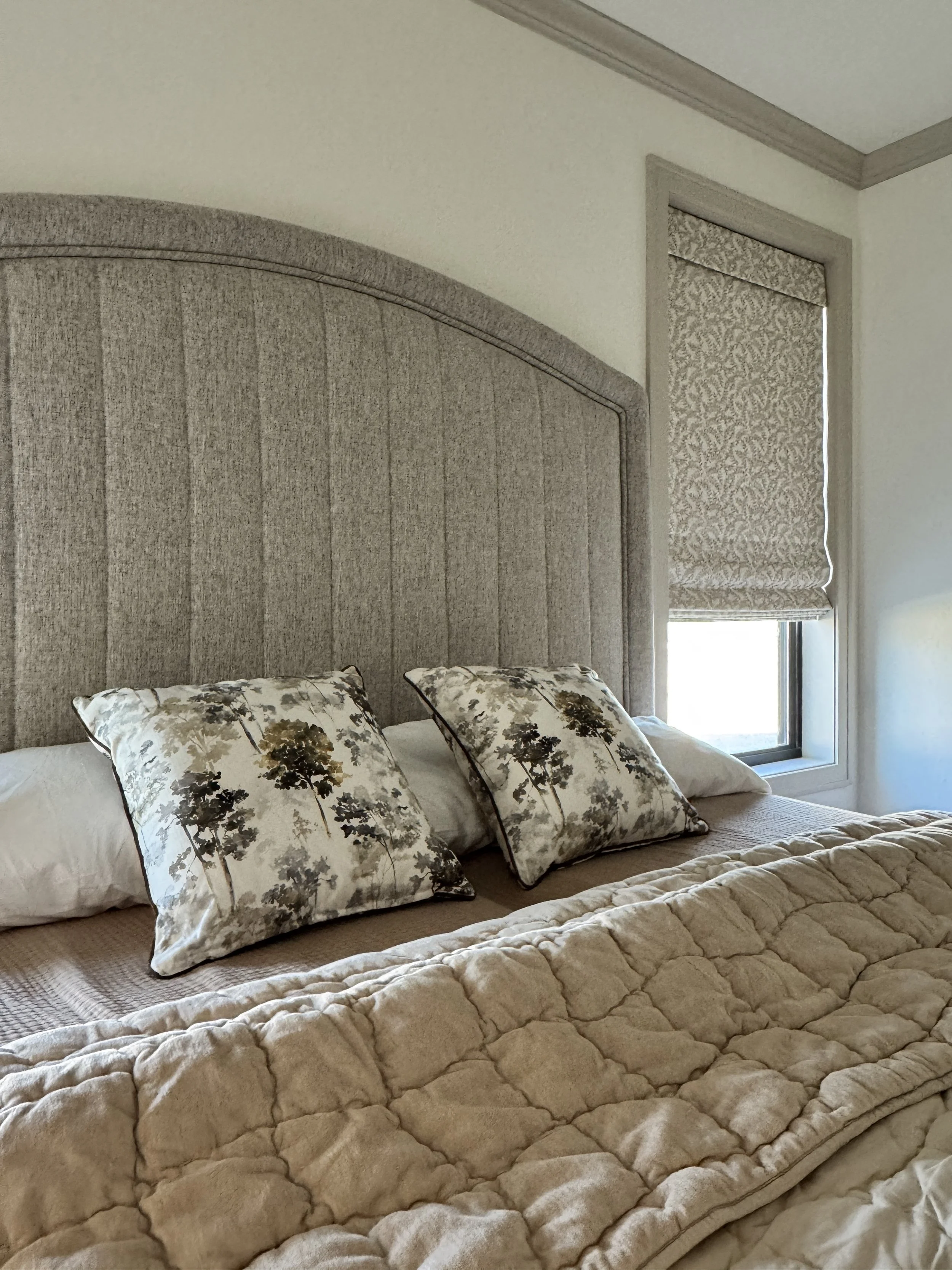 A bedroom with a large, upholstered headboard, two decorative pillows with floral patterns, a quilted blanket, and a window with a patterned roller blind.