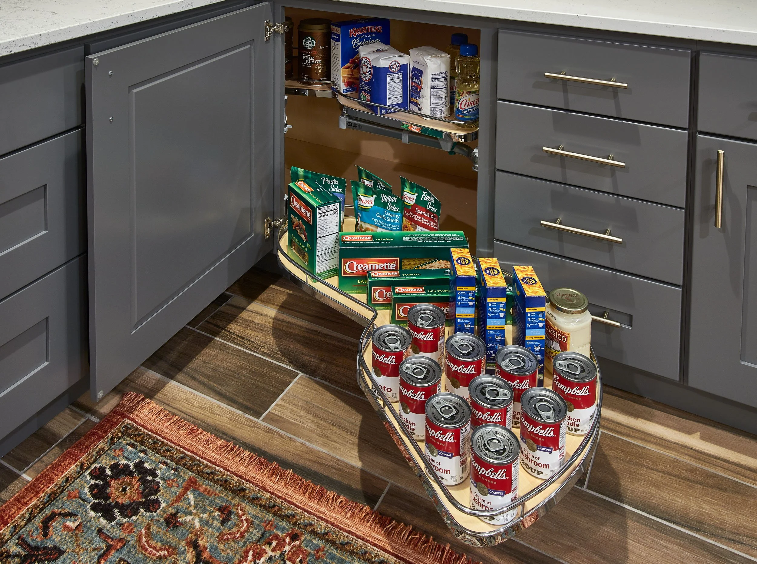 Open kitchen cabinet with food items like canned soup, pasta, and spices, with pull-out shelves showing canned soup in the foreground and boxed pasta and spices in the background.
