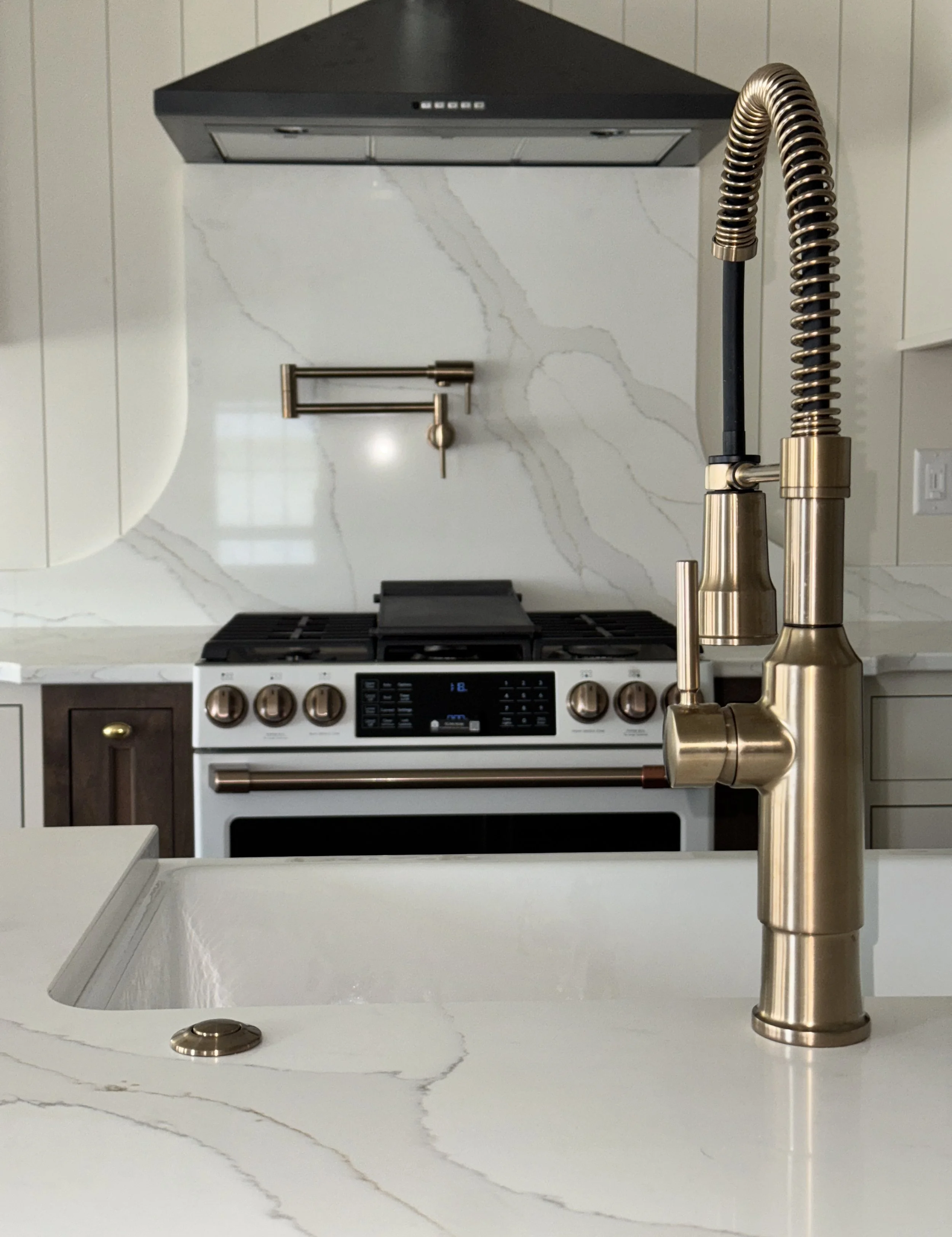 Close-up of a modern kitchen sink with a gold faucet in front of a white stove with gold knobs, and a marble backsplash and countertop.