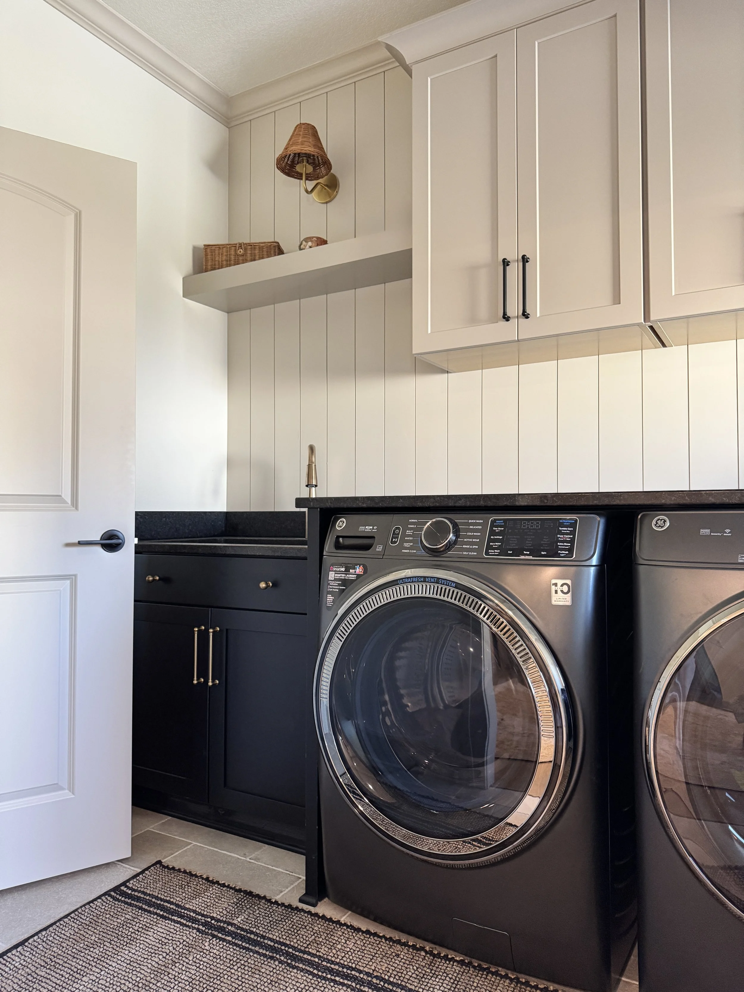 A laundry room with magnetic navy blue cabinets, beige wall paneling, a white door, a black countertop with a sink, a silver front-loading washing machine, and a beige rug on the tiled floor.