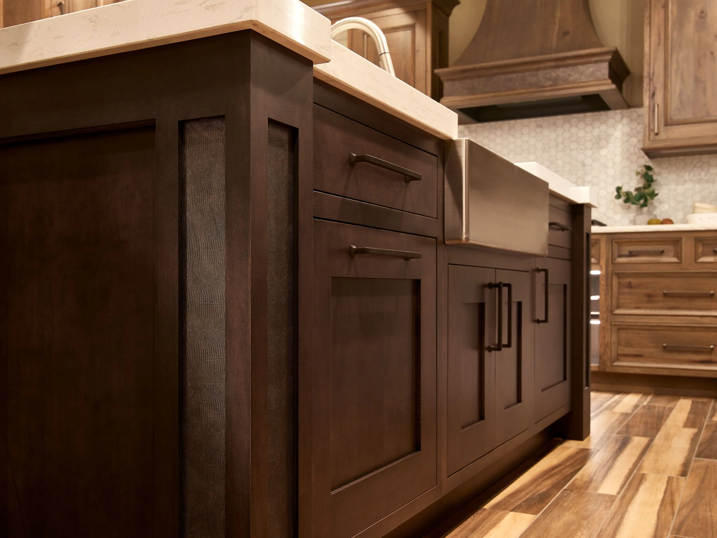 Close-up of a kitchen island with dark brown wood cabinetry, a white countertop, and an undermount stainless steel sink. In the background are matching wooden kitchen cabinets and drawers, with a hexagonal tile backsplash and a small plant.