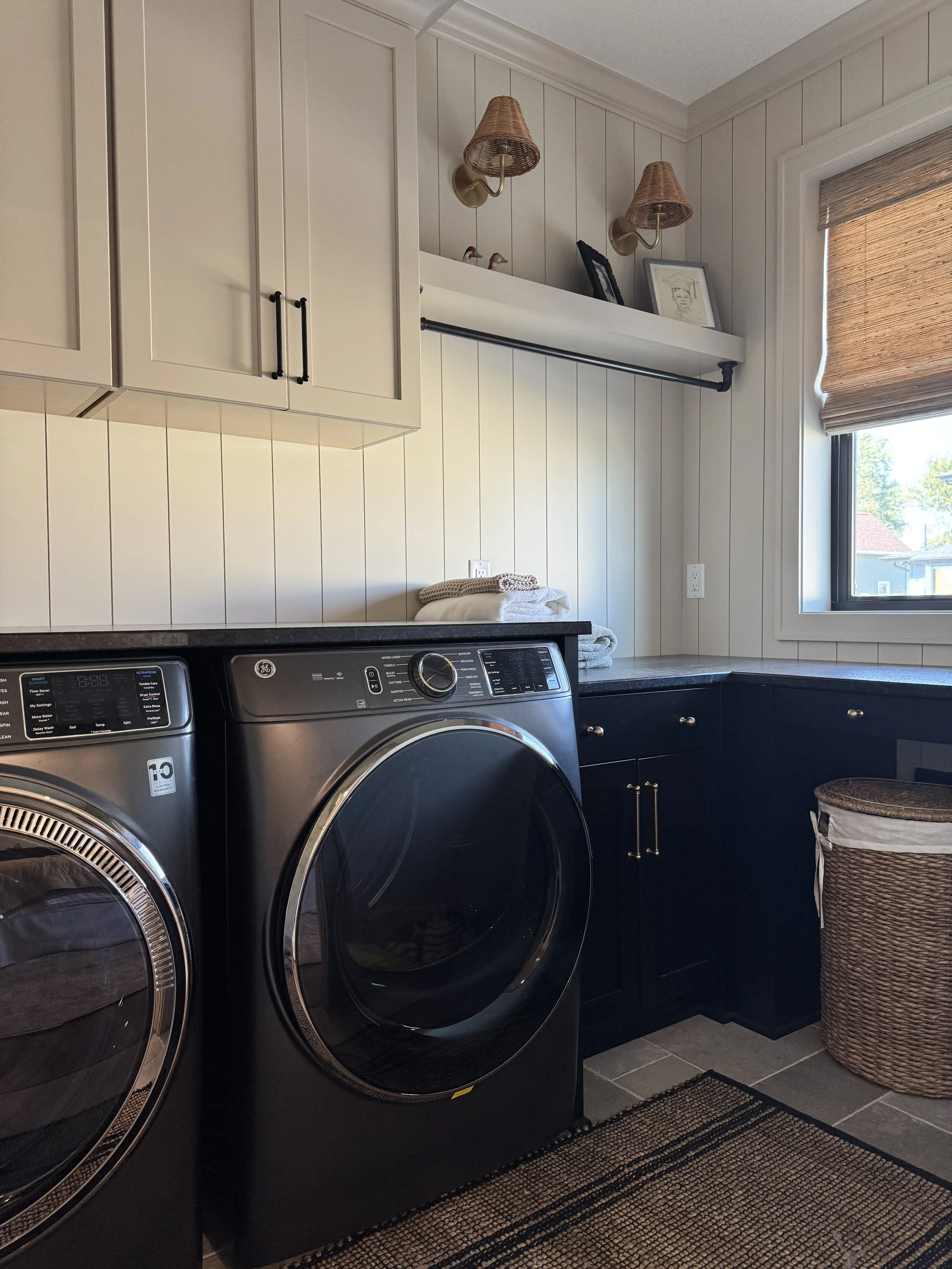 Laundry room with white cabinets, black washer and dryer, window with brown blinds, wicker basket, and decorative wall lamps.