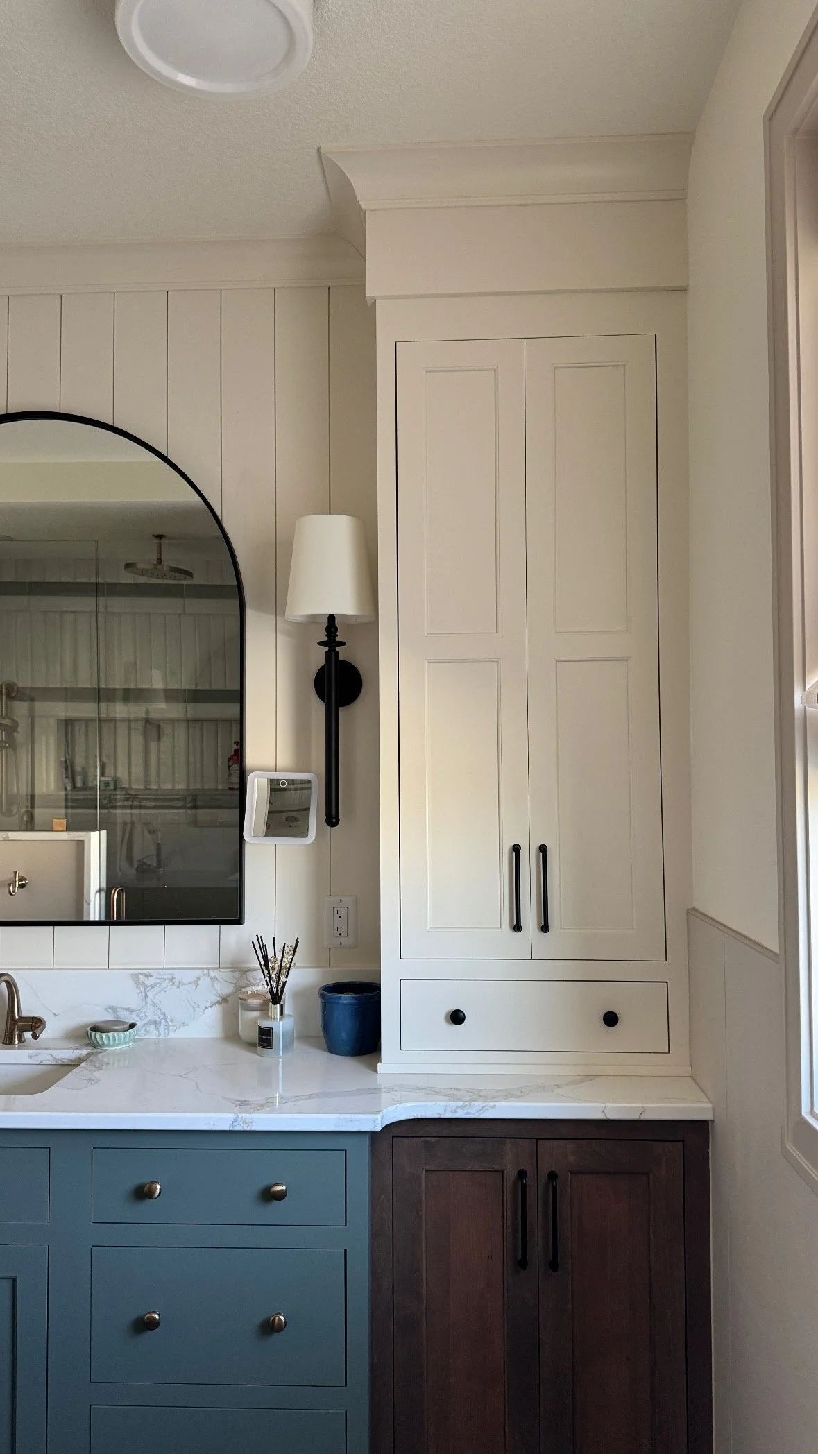 A kitchen or bathroom area with a white marble countertop, blue cabinets, and a tall white cabinet. There is a black wall sconce with a white lamp shade, a mirror with an arched top, and a small square device on the wall. A blue pot, a reed diffuser,