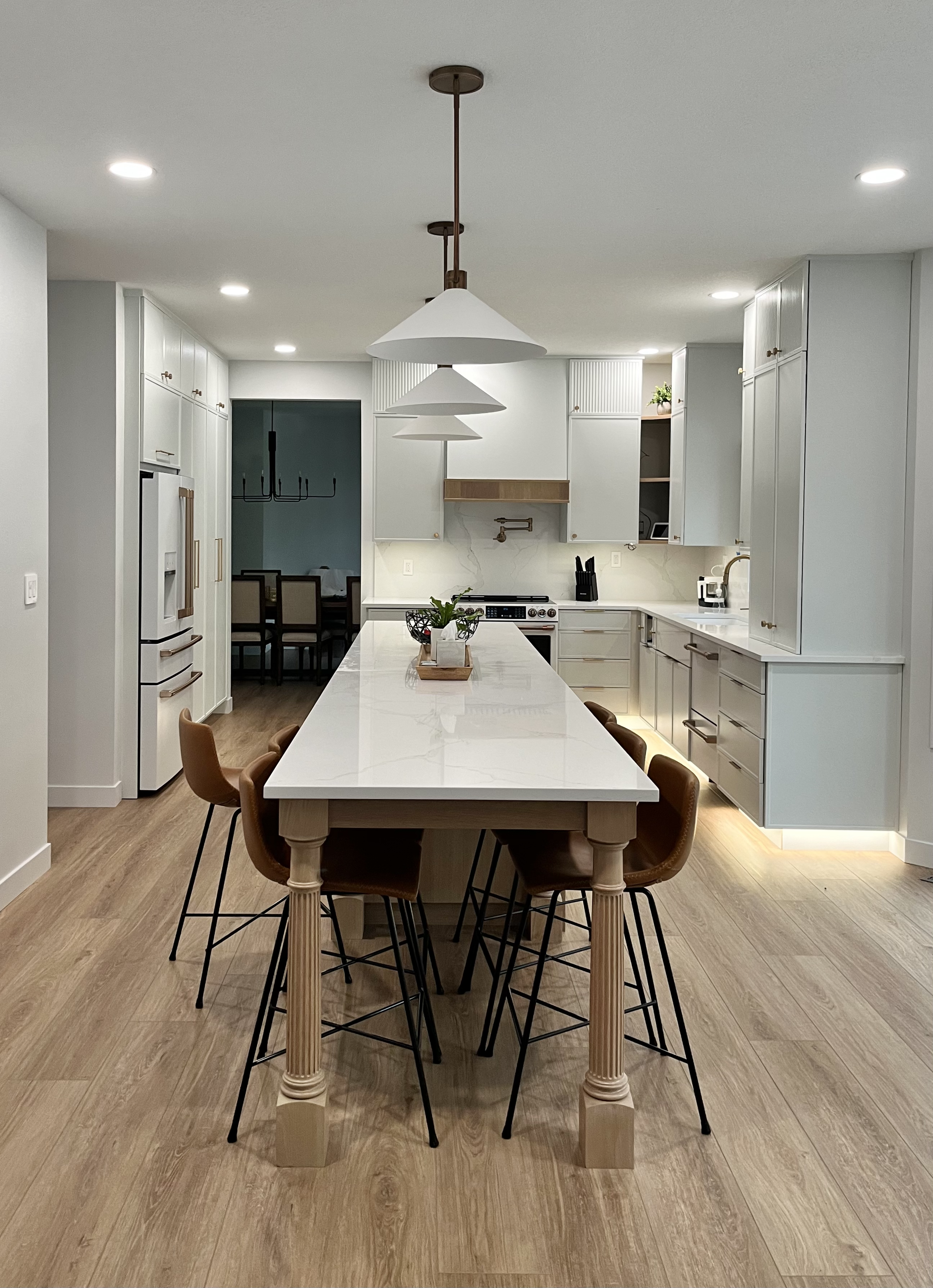 Modern kitchen with an island table, brown chairs, white cabinets, and wood flooring. There are three pendant lights hanging over the island and some recessed lighting in the ceiling. A small plant is on the island.