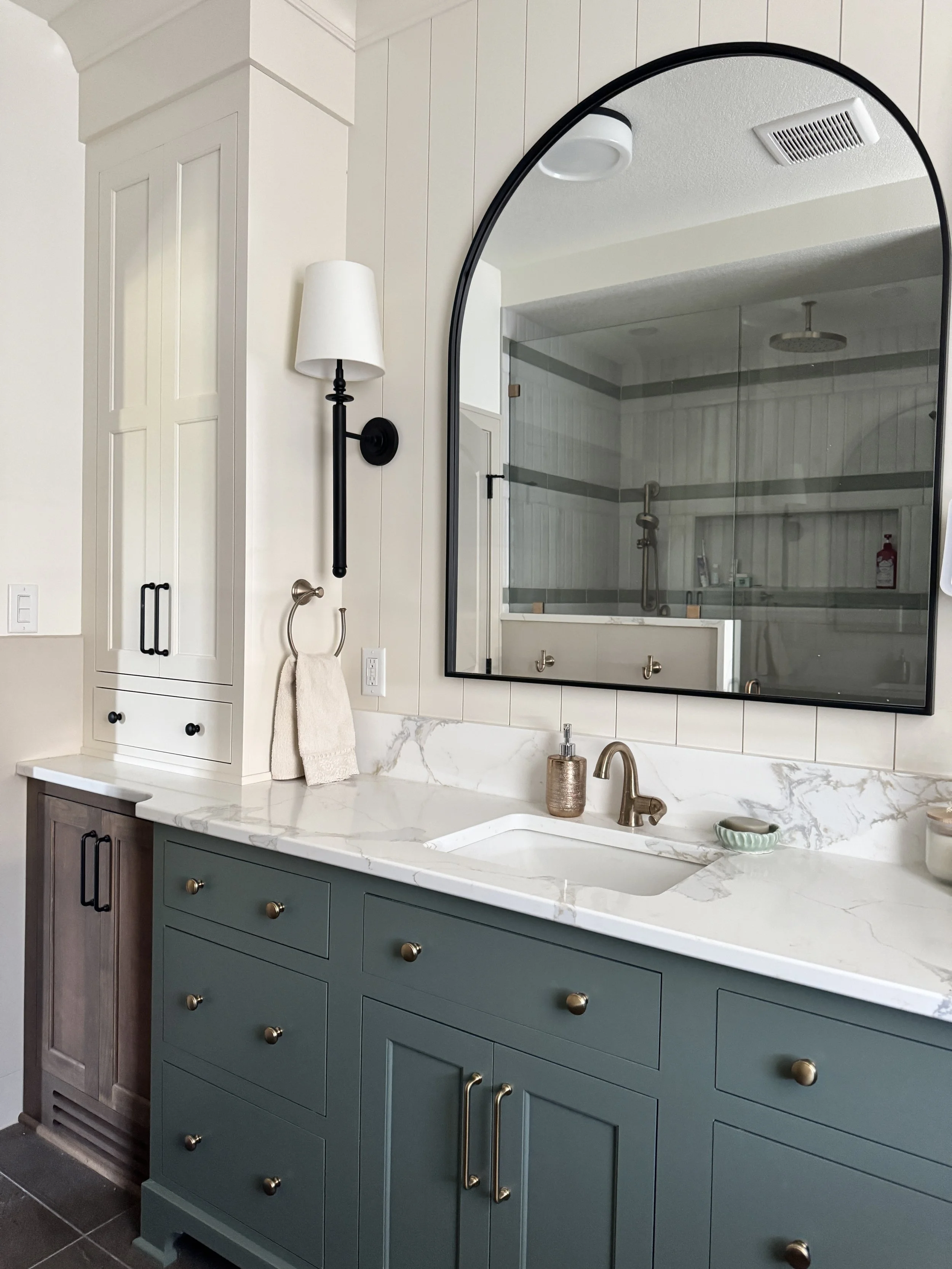 Bathroom vanity with green cabinets and marble countertop, large mirror with black frame, brass faucet, soap dispenser, and small decorative bowl, with shower visible in reflection.