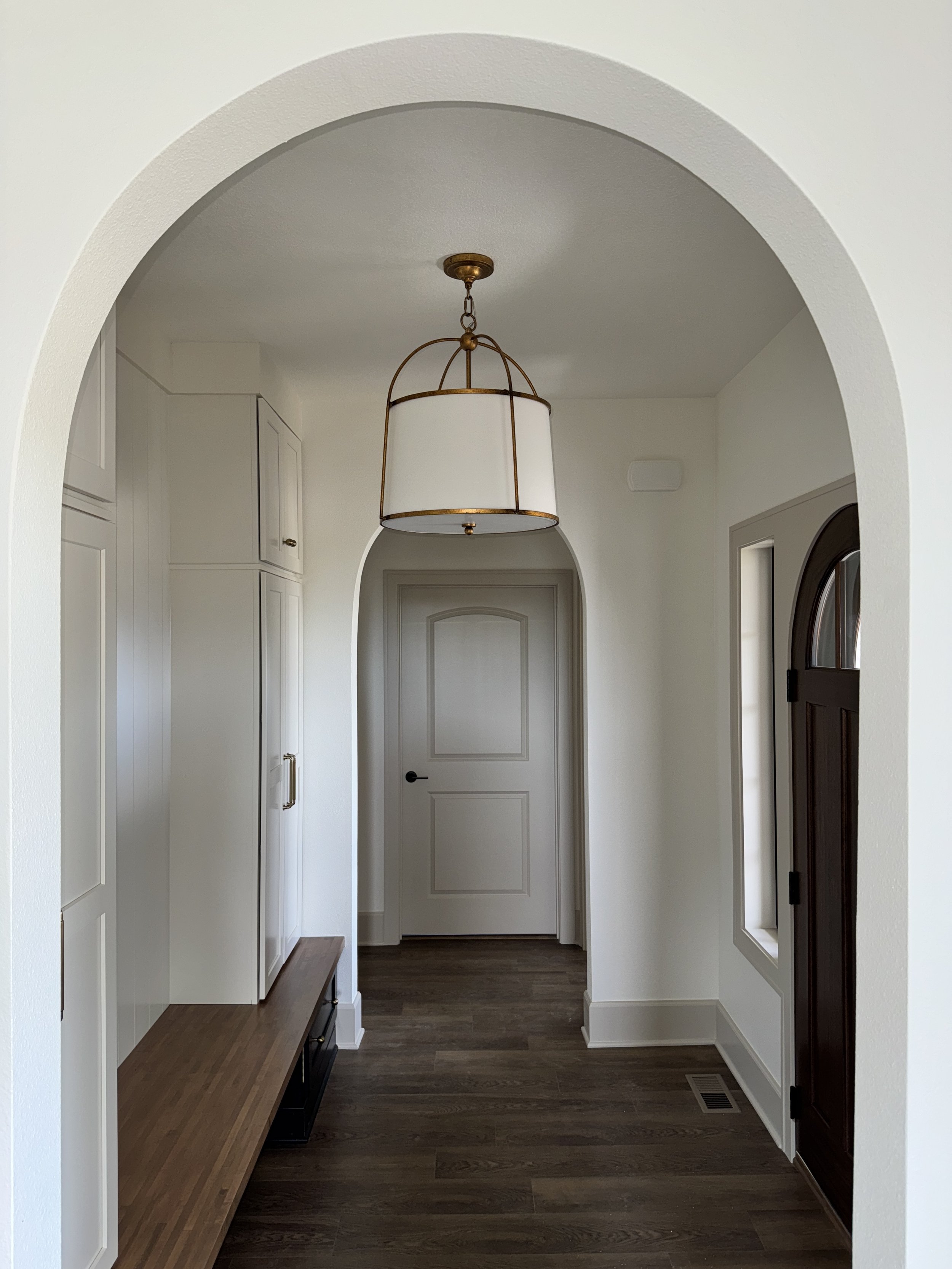 Entryway with white walls, wooden floor, a white door at the end, a decorative light fixture hanging from the ceiling, and a bench on the left side.