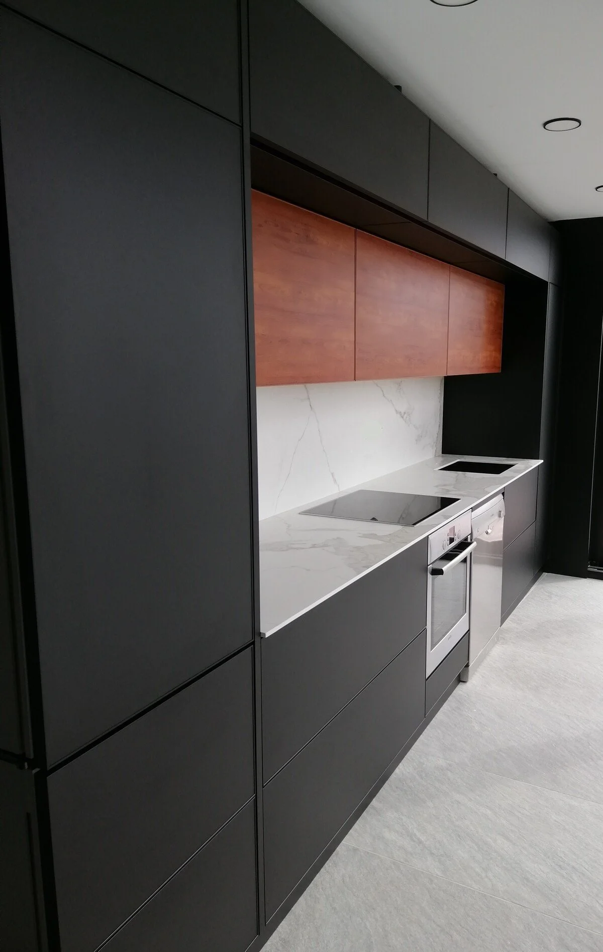 Modern kitchen with black cabinetry, white marble countertop, and wooden upper cabinets, featuring an electric cooktop and oven.