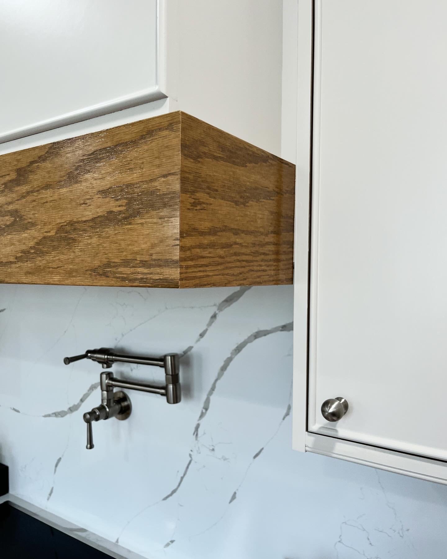 Close-up of a kitchen corner showing a wooden cabinet and white marble backsplash with gray veins, and a dark metal faucet.