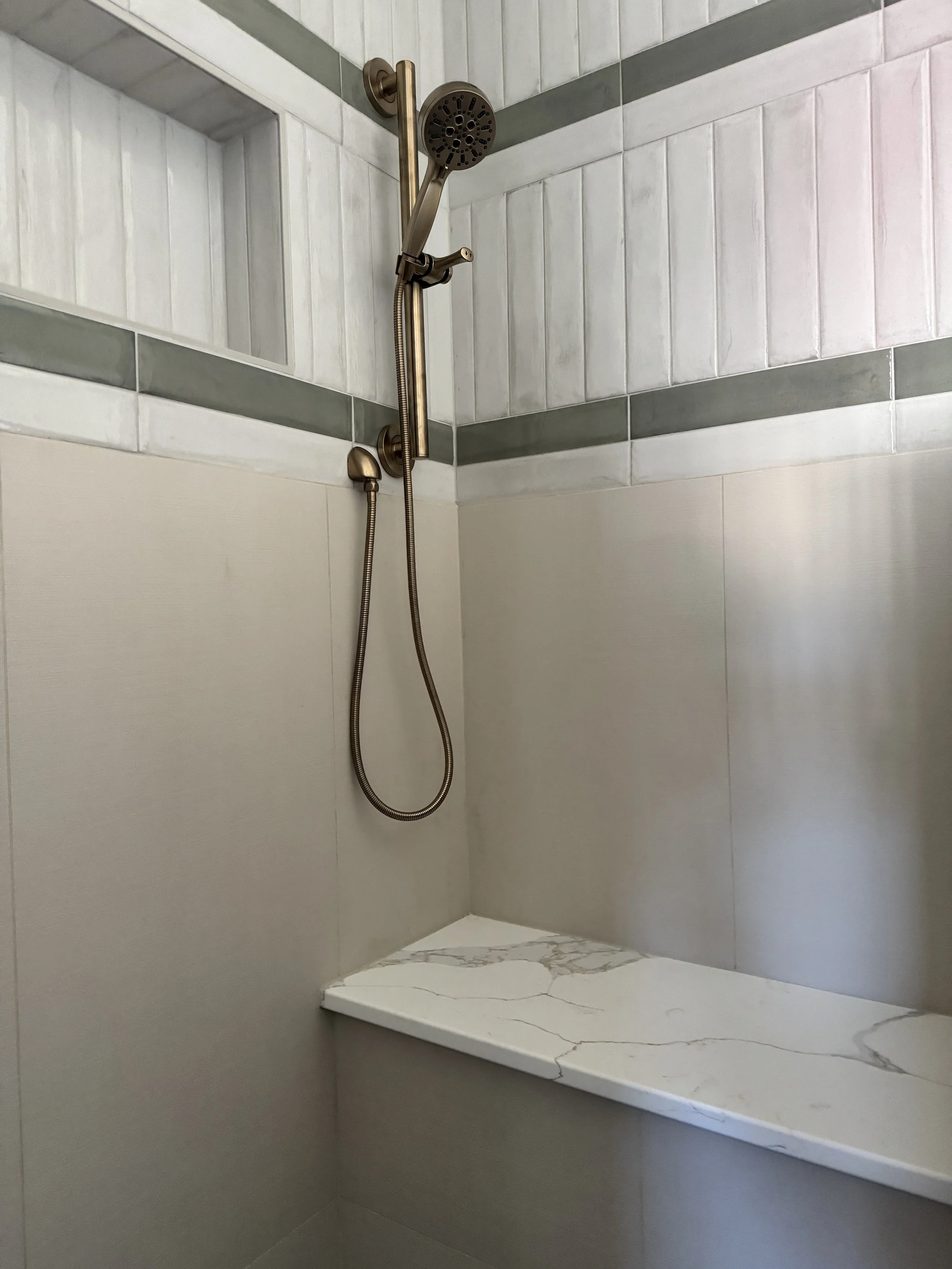 A corner shower with a bronze showerhead, a movable handheld shower, white and gray tiled walls, and a marble-like bench.
