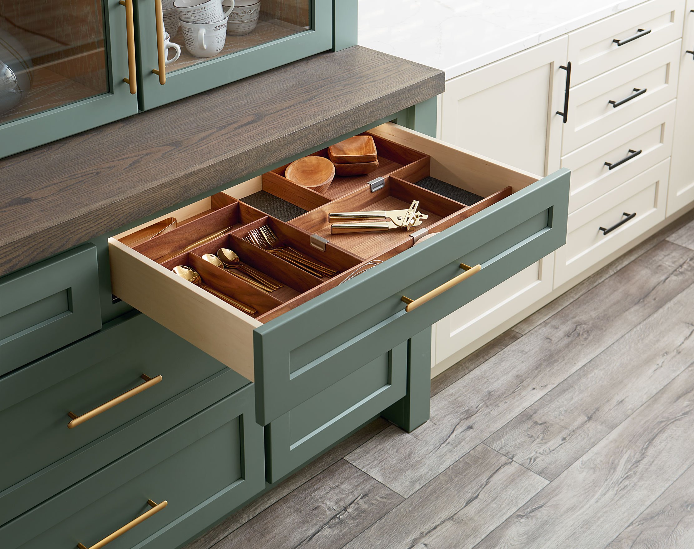Open kitchen drawer with wooden utensils and cutlery, nearest green cabinetry and white cabinetry, wood flooring.