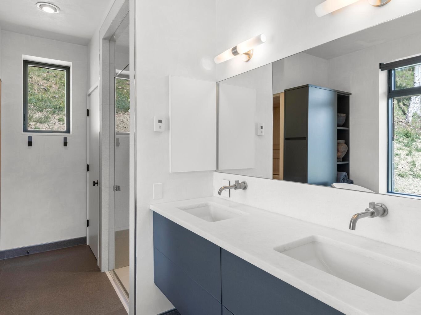 Modern bathroom with double white sinks, large mirror, natural light from windows, gray and white color scheme, and minimalist decor.