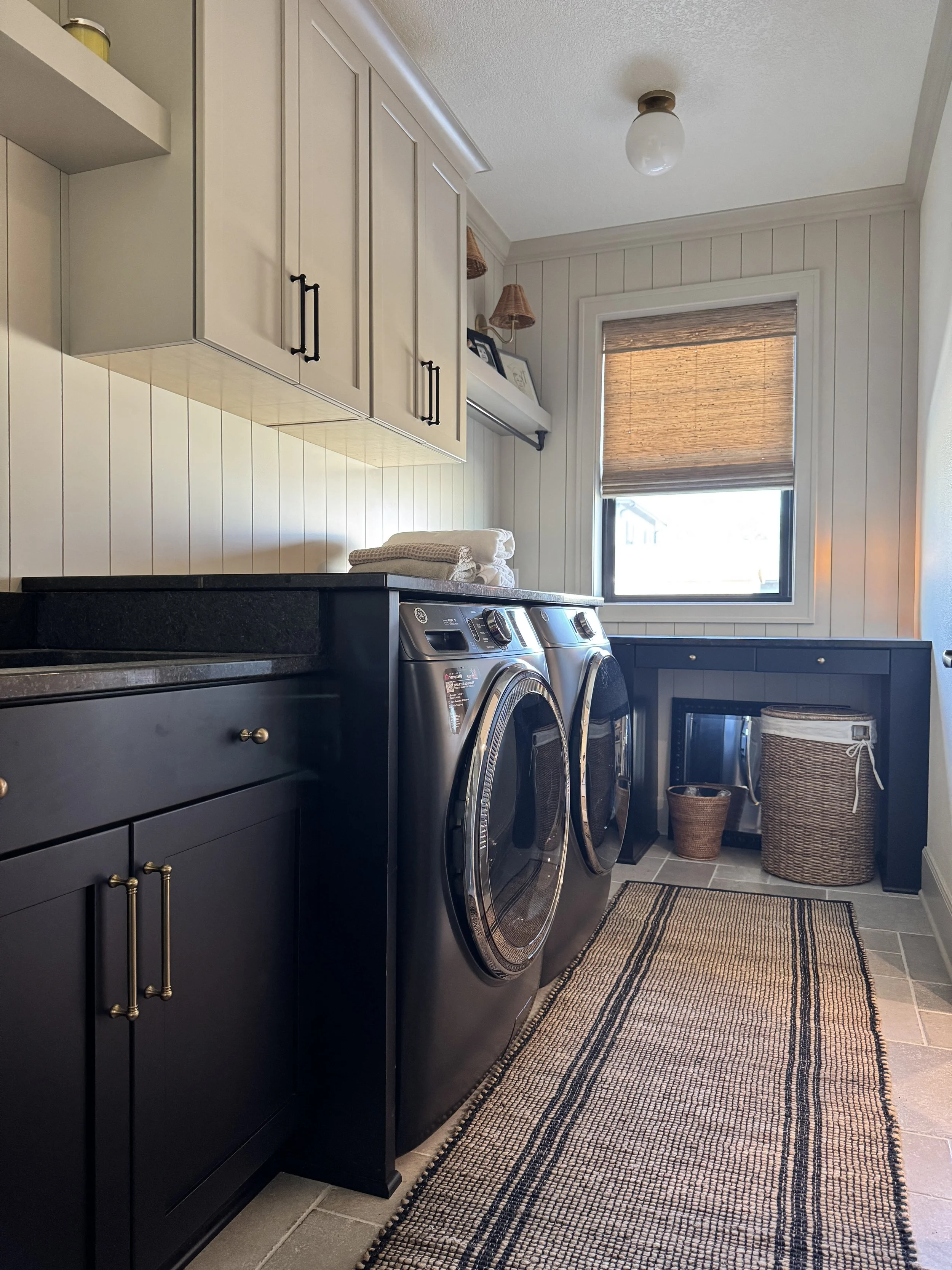 A laundry room with black washer and dryer, white cabinets, window with a brown blind, wicker basket, and beige wall paneling.