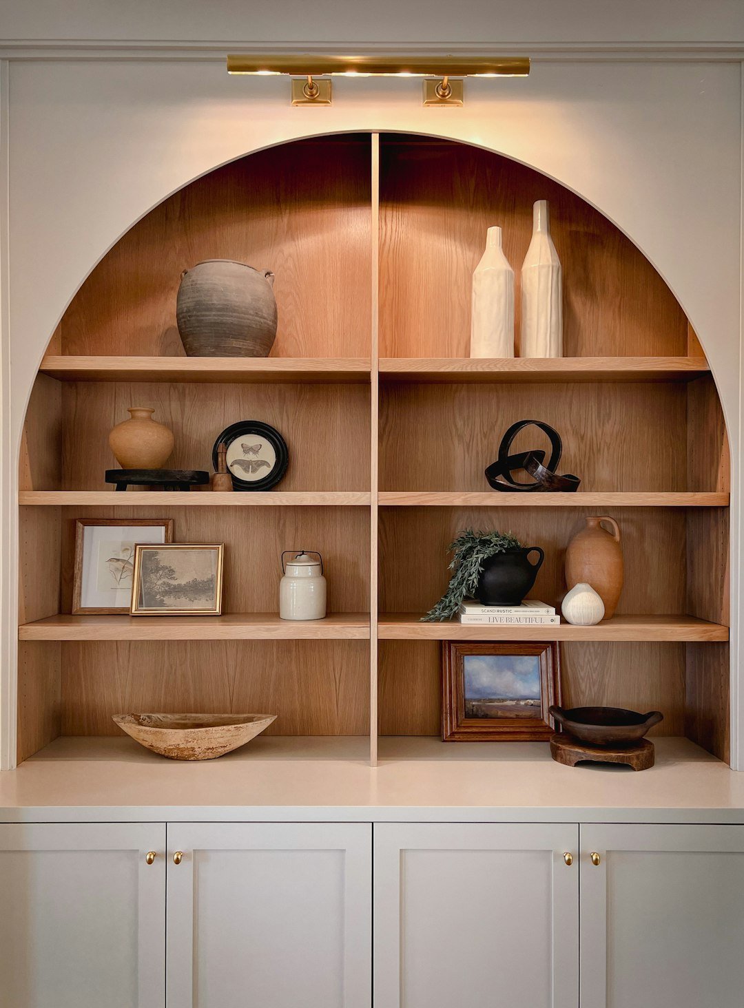 A laundry room with black washer and dryer, white cabinets, a window with a bamboo shade, a woven basket, and a shelf with framed photos and decorative items.