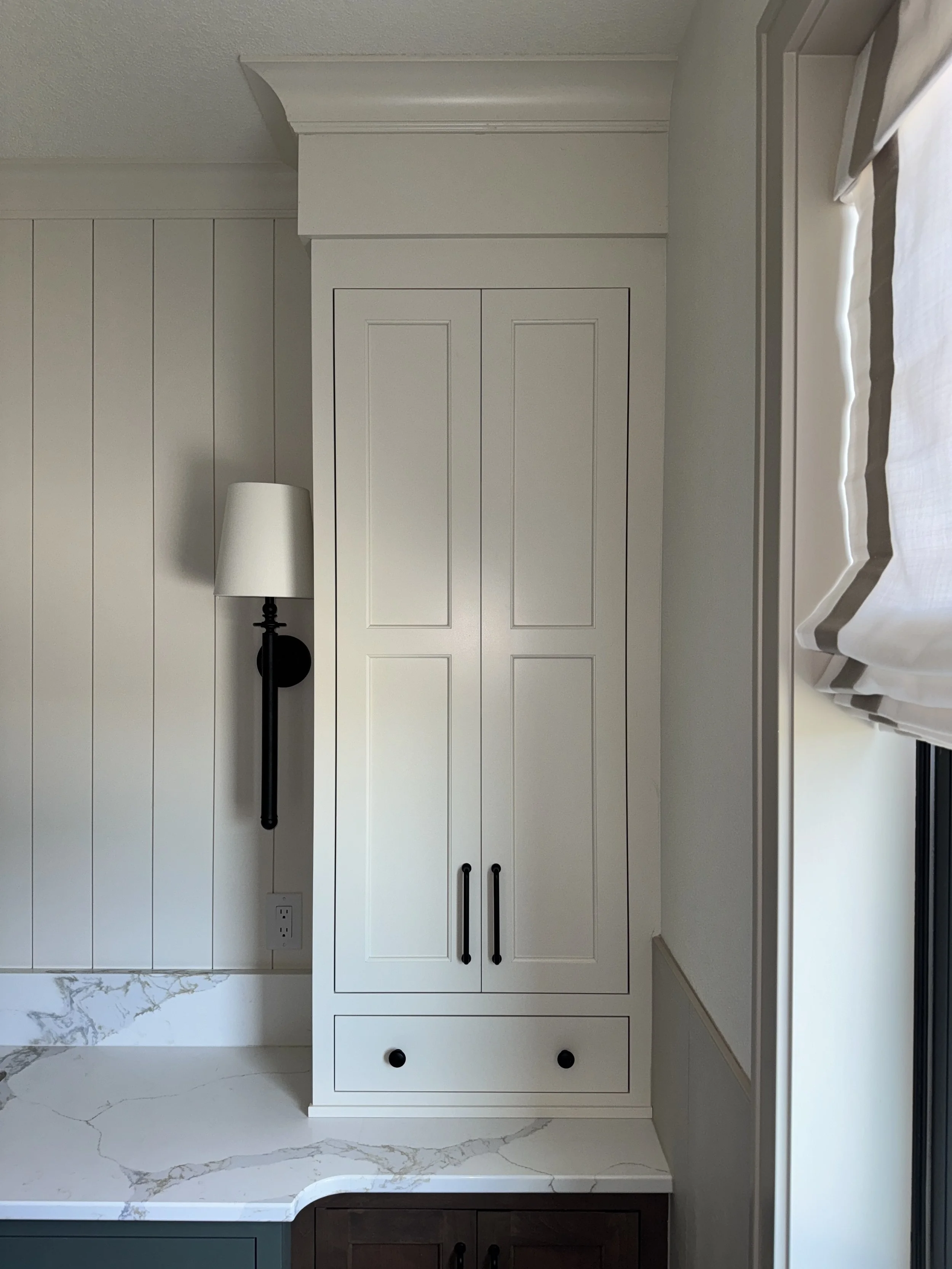 A white kitchen cabinet with black handles and two black knobs at the bottom, next to a white marble countertop, with a black wall-mounted lamp and a window with white shades.