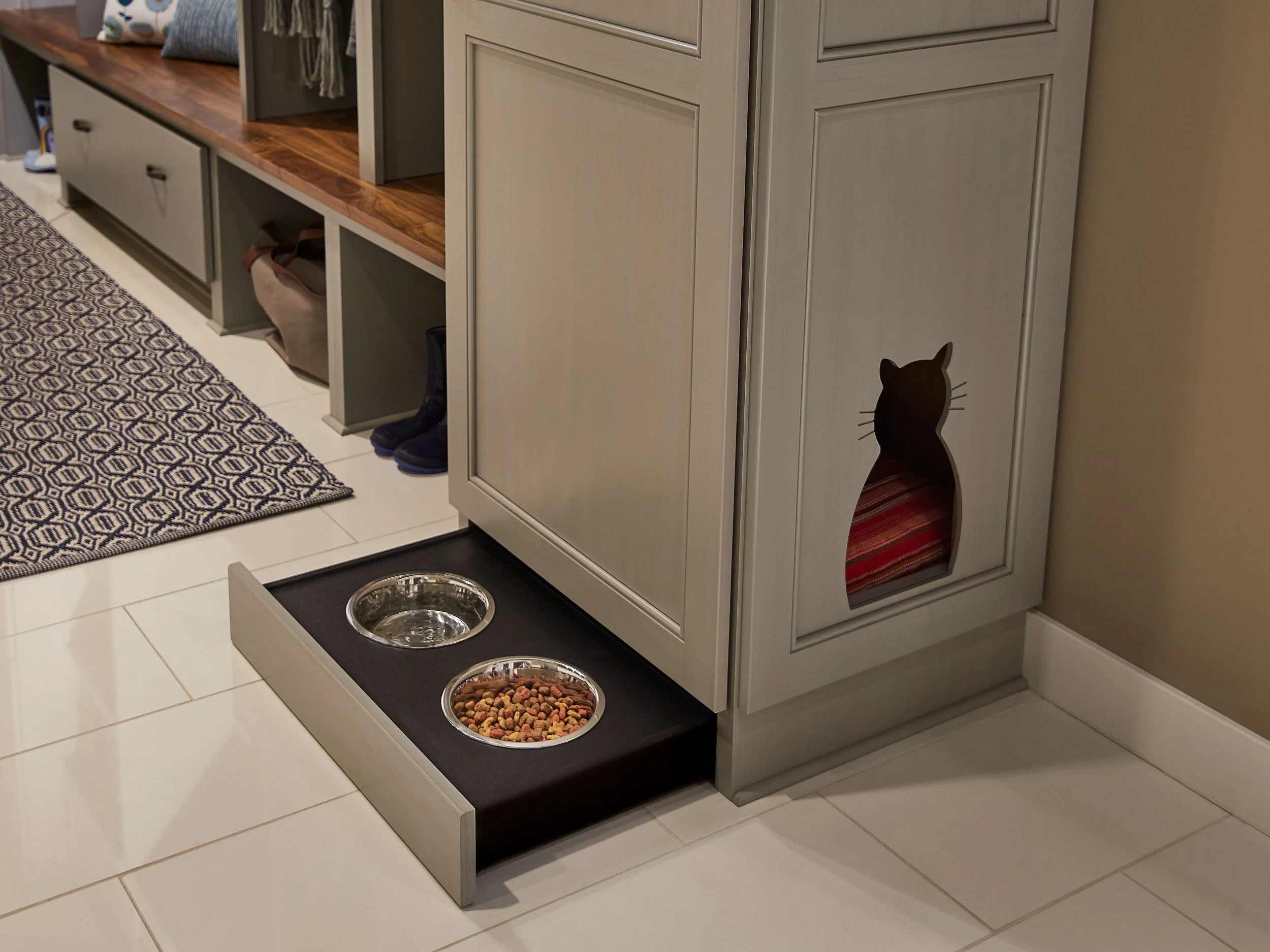 A cat-shaped cutout in a cabinet door, with a blanket inside. Two metal bowls in a black tray, one with water and the other with dry cat food, are on the floor nearby.