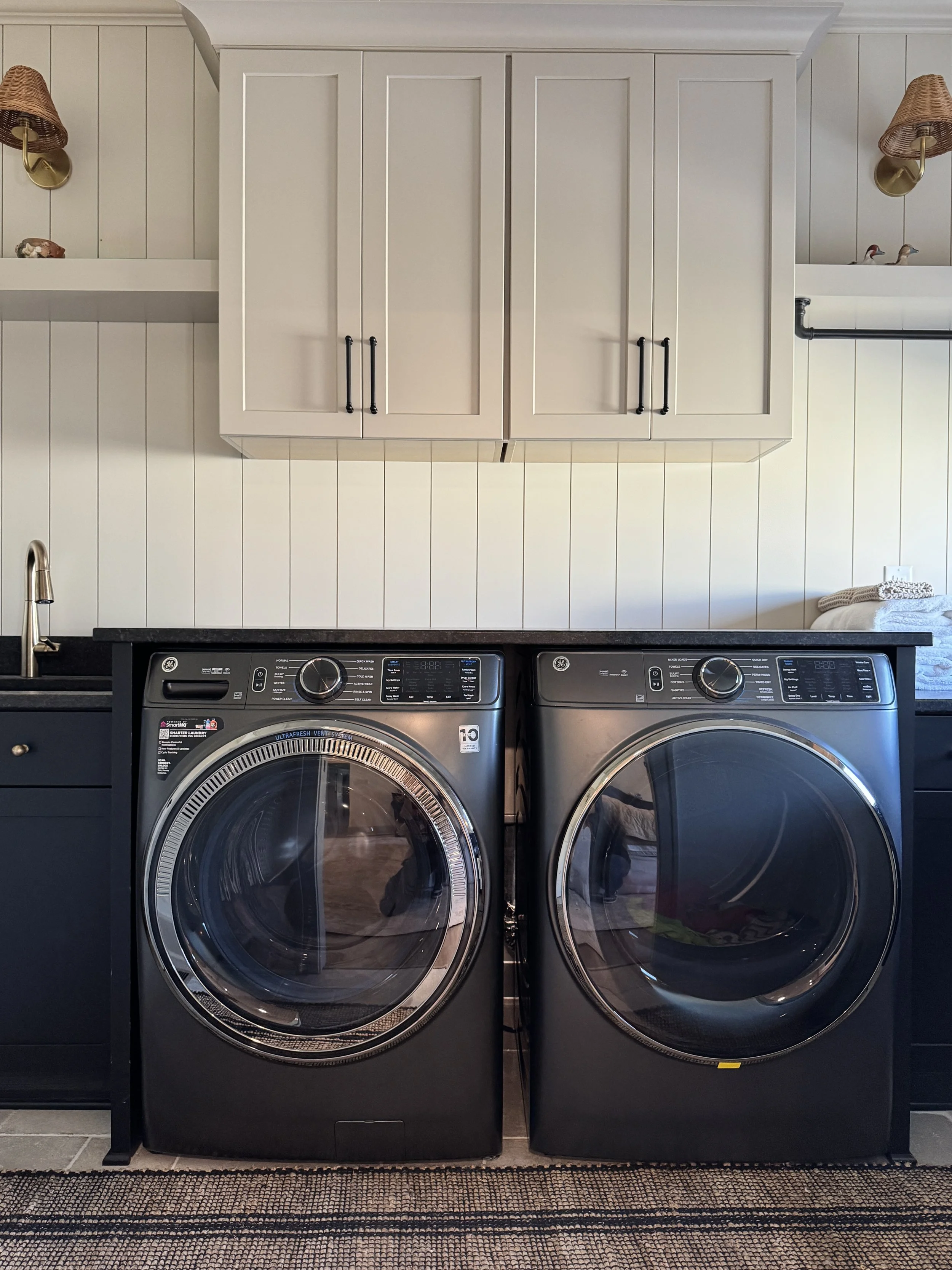 Front view of a laundry area with a black washer and dryer side by side, wooden cabinets above, a white wall with vertical paneling, and wall-mounted lamps on each side.