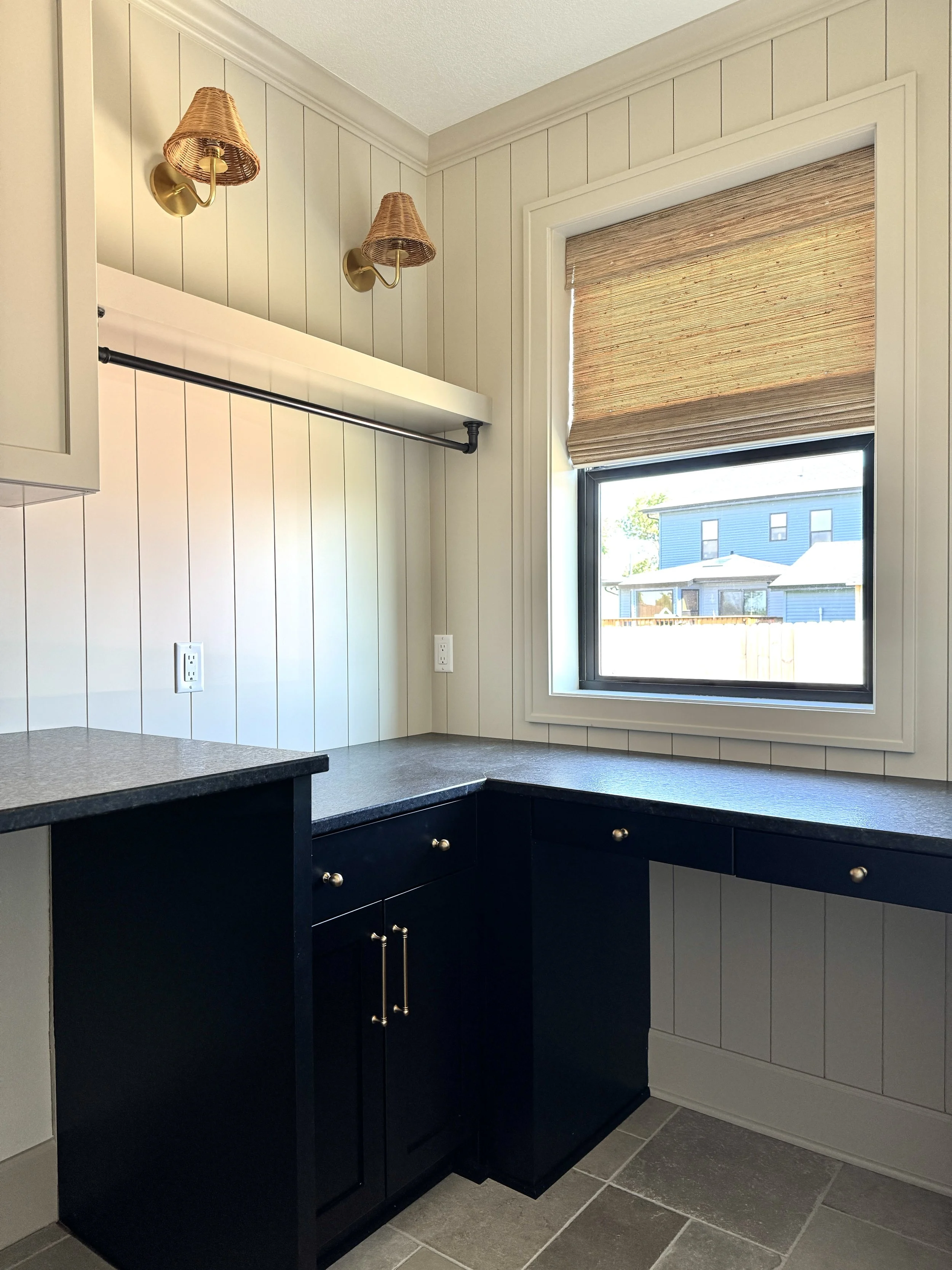 Corner of a room with white paneled walls, a window with a brown woven shade, black countertop, black cabinets with gold handles, two brass wall sconces with wicker lampshades, and tiled flooring.