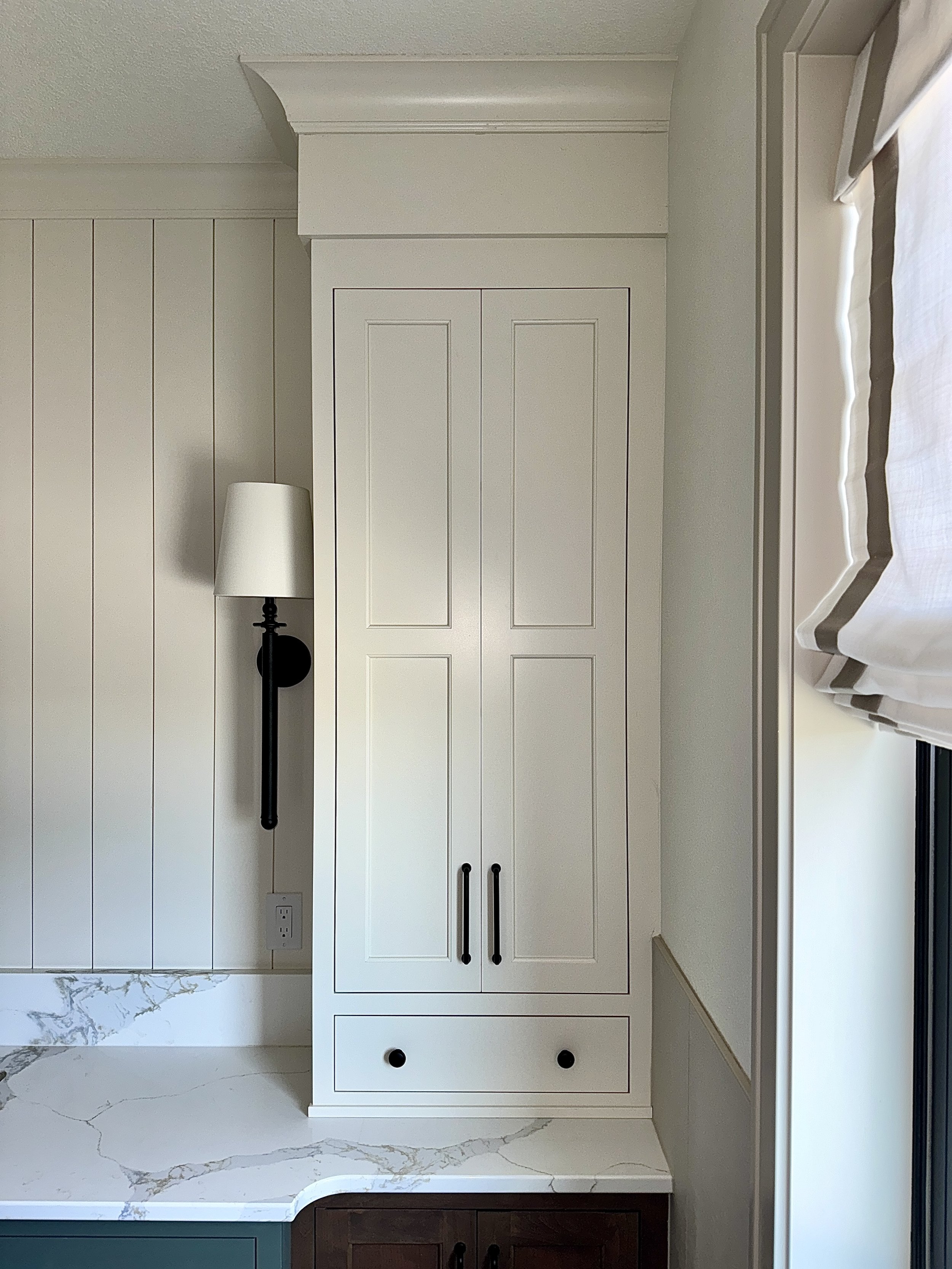 White built-in kitchen cabinet next to a window with white Roman shades, a black wall-mounted lamp with a white lampshade, and a marble countertop with a dark wooden cabinet below.