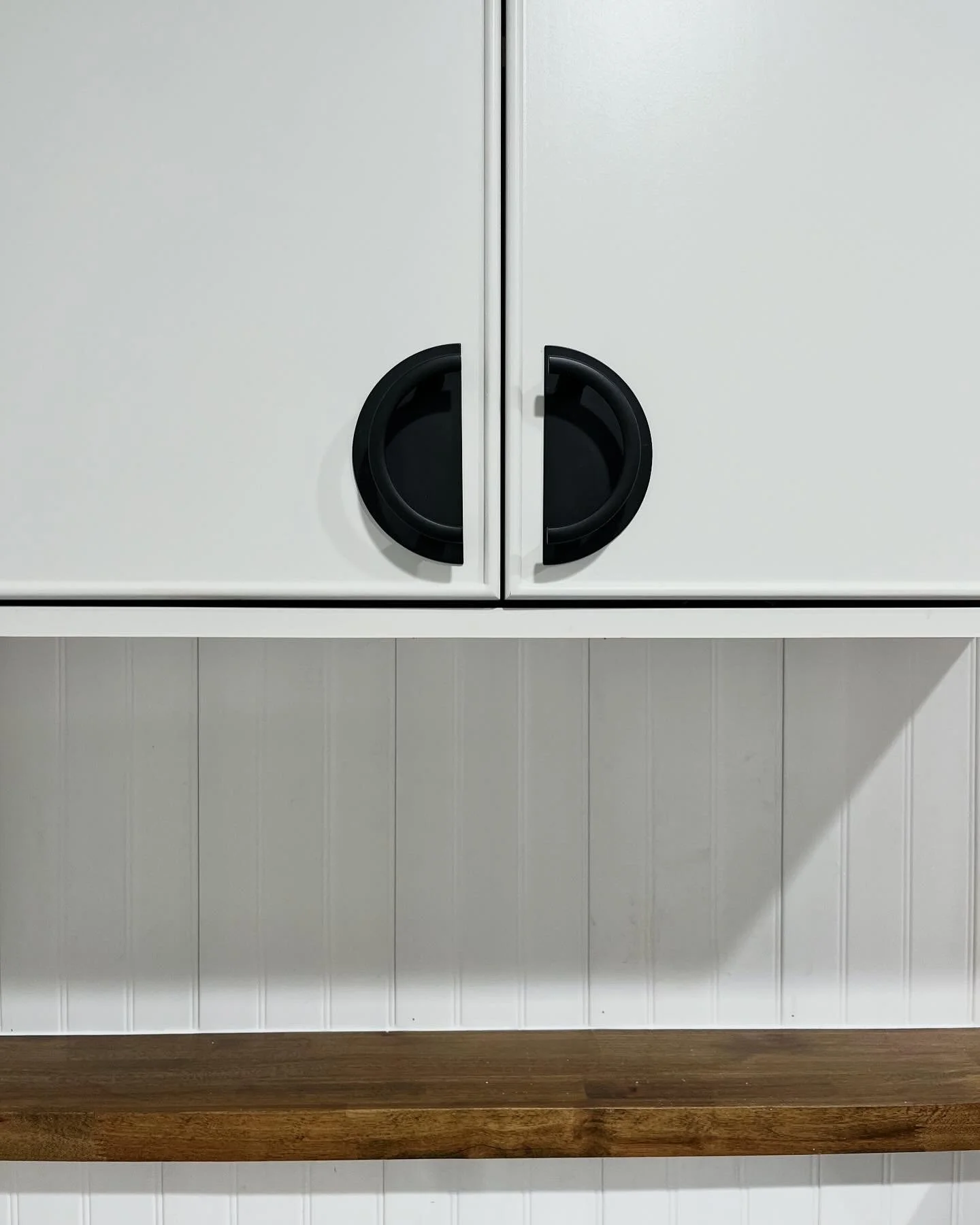 Close-up of a white cabinet with black circular handles, mounted on a wall with vertical white paneling, and a wooden shelf below.