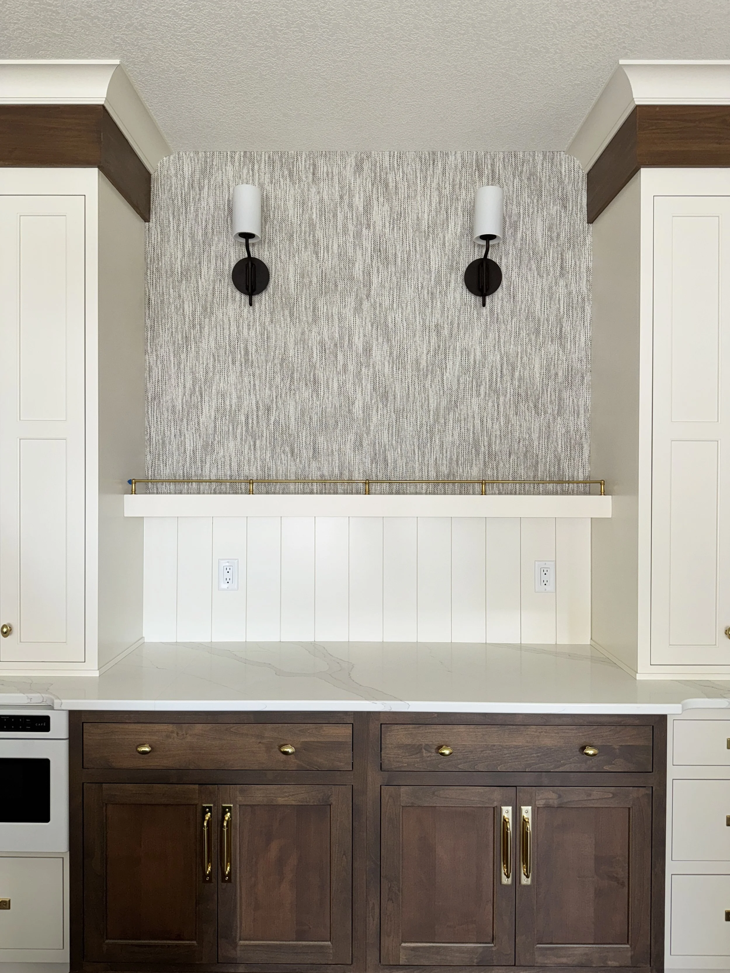 A kitchen countertop with white and dark wood cabinets, two wall sconces with white shades, and a textured wall with vertical gray and white pattern.